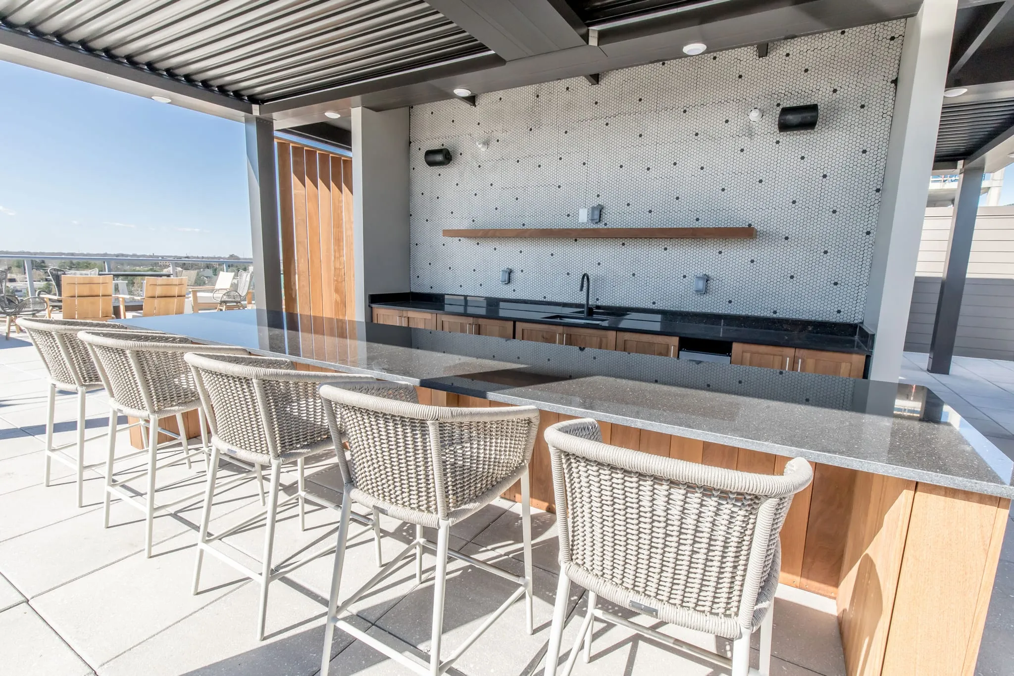Multifamily amenity rooftop bar with woven bar stools along the counter under the pergola, the city beyond