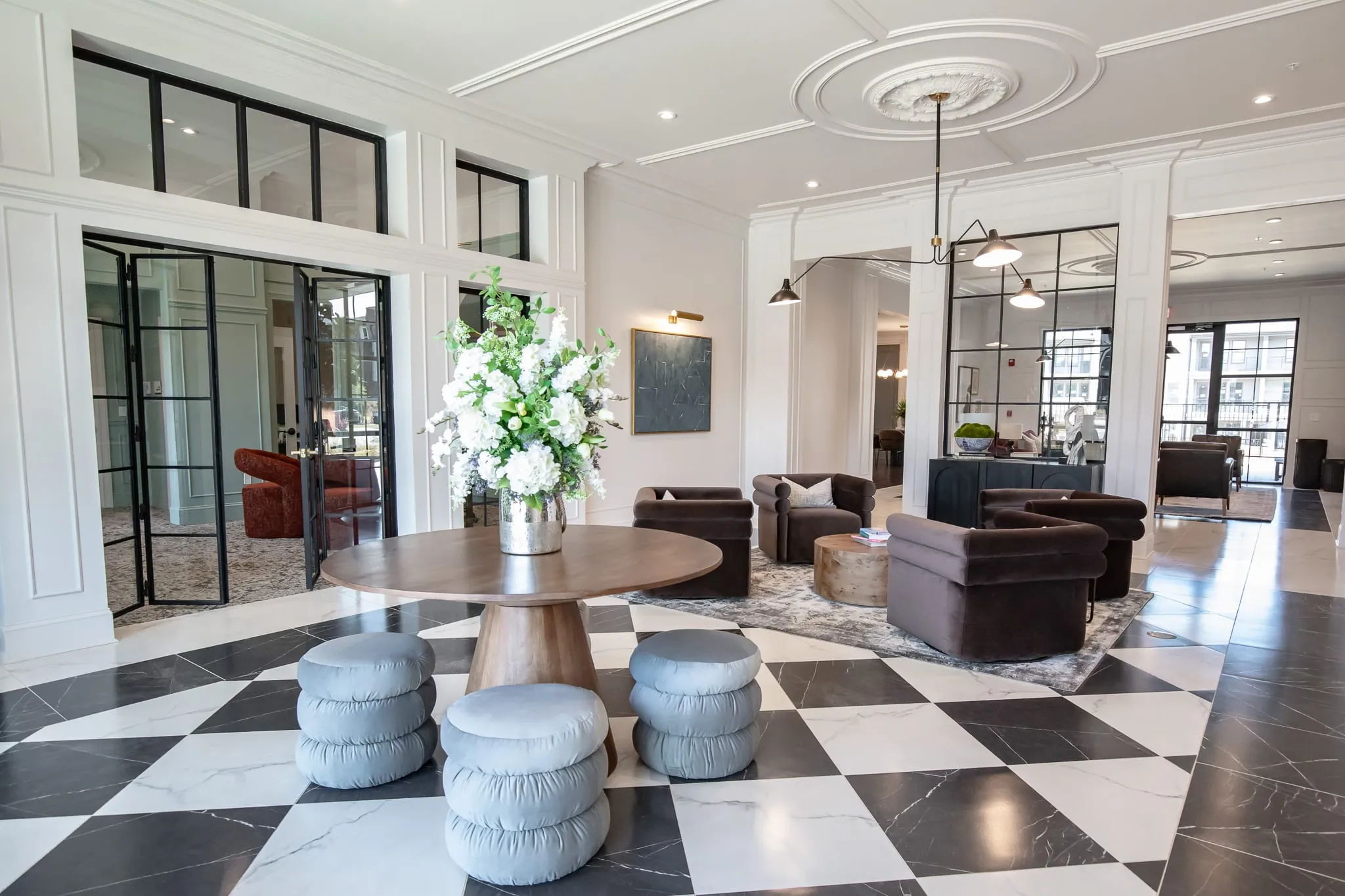 Apartment lobby with a black-and-white chequerboard marble floor, a round wood pedestal table holding white hydrangeas, grey tufted pouf stools and brown velvet swivel chairs under a coffered ceiling