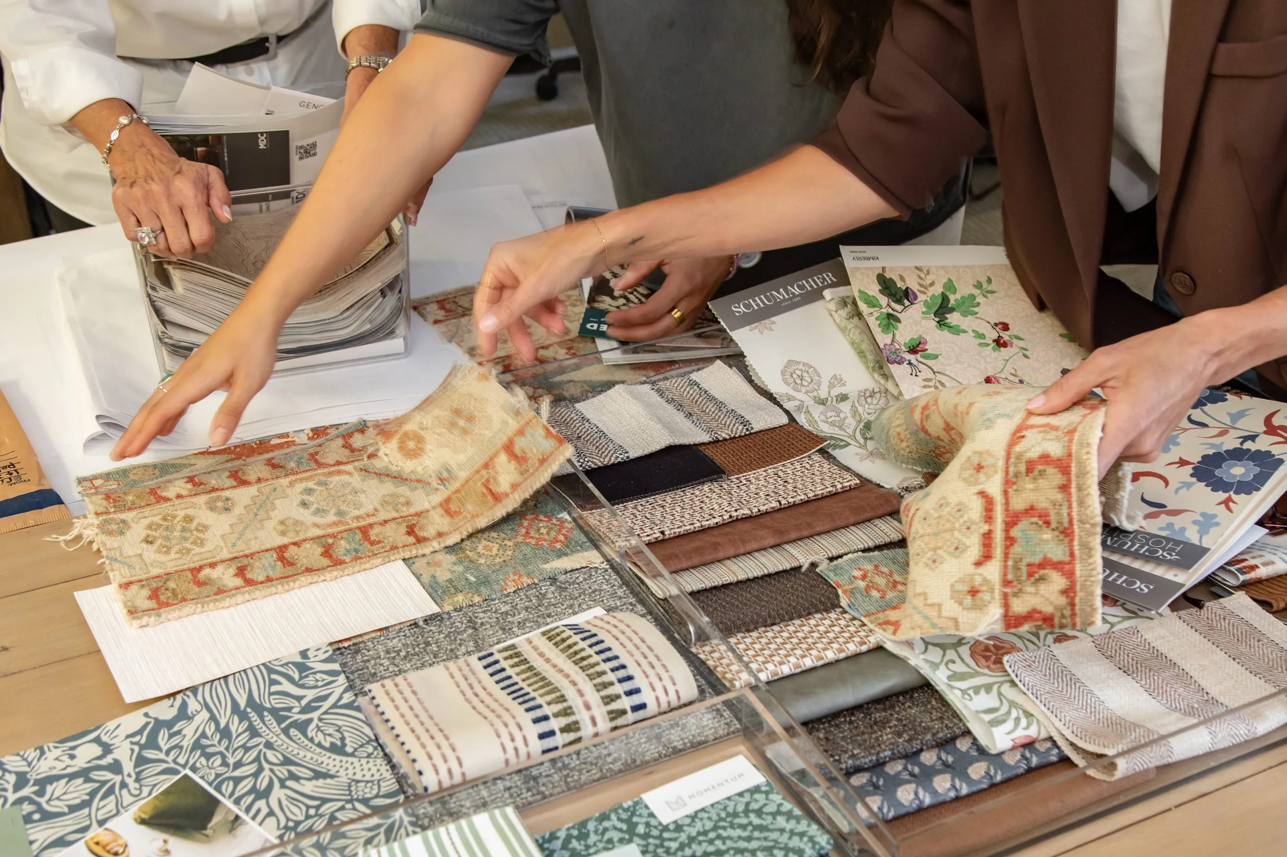 Overhead view of three designers' hands sorting rug swatches, striped fabric samples and wallpaper books spread across a table