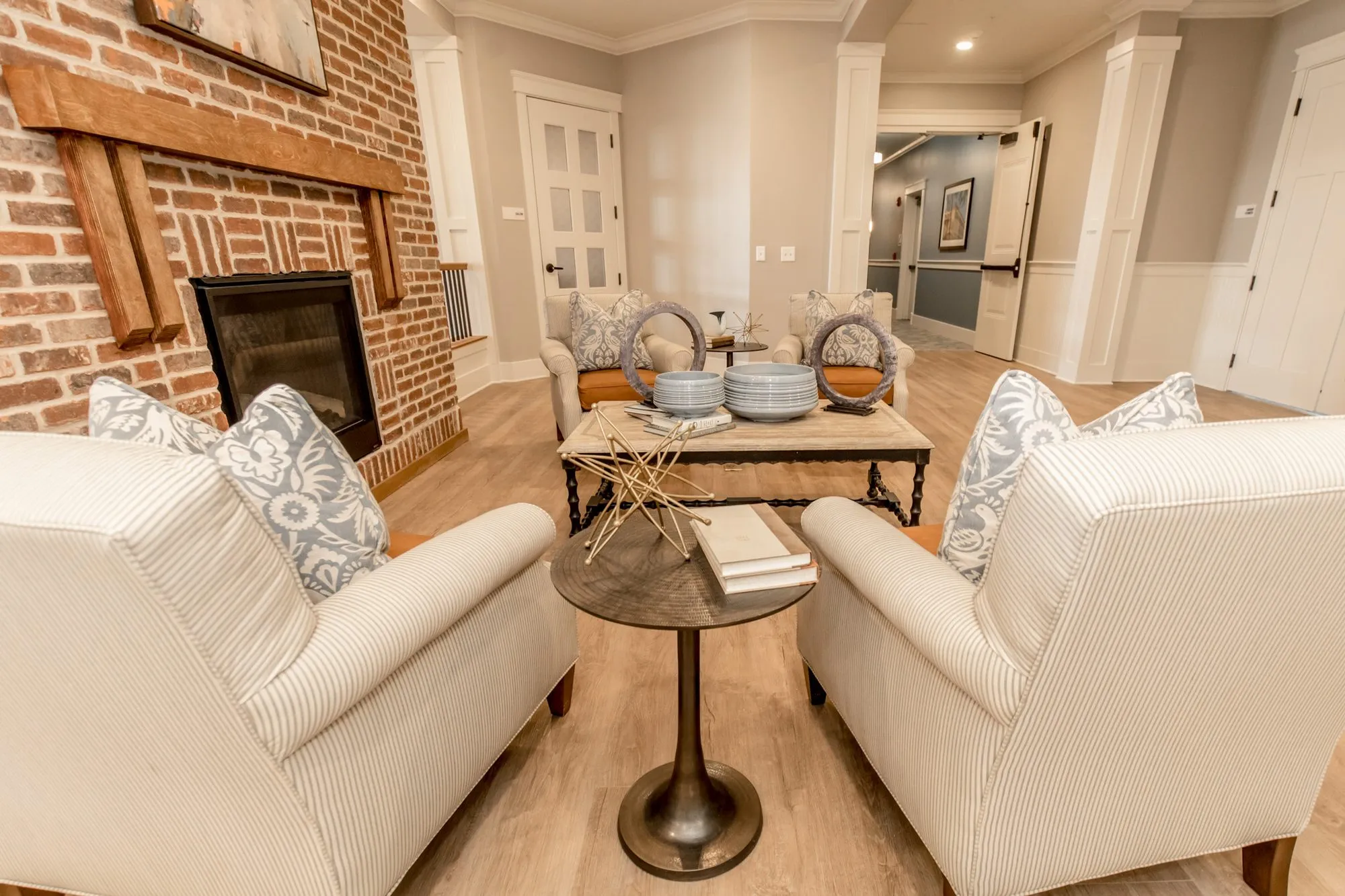 Senior living lounge with striped armchairs at a bronze side table by the brick fireplace and timber mantel, corridor beyond