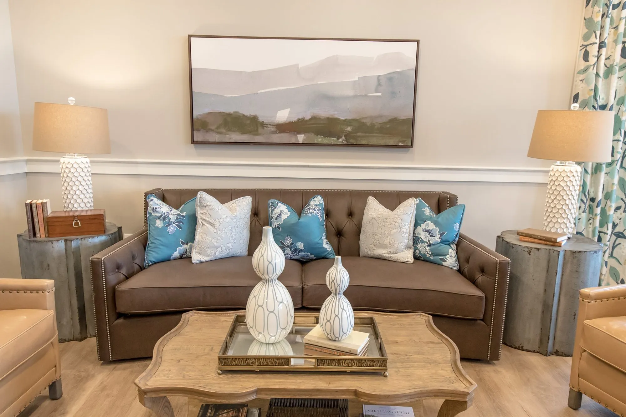 Senior living interior detail: brown tufted sofa with blue floral pillows below a landscape painting, lamps on drum tables