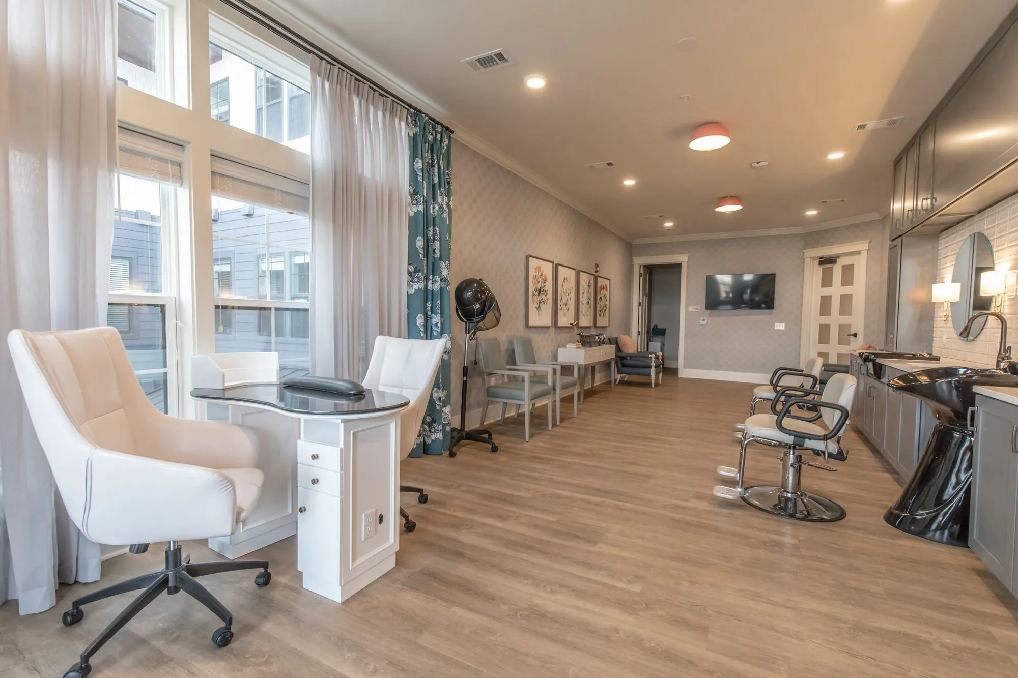 Senior living salon with a manicure table and white chairs by tall windows, a styling chair and wash basins along the wall
