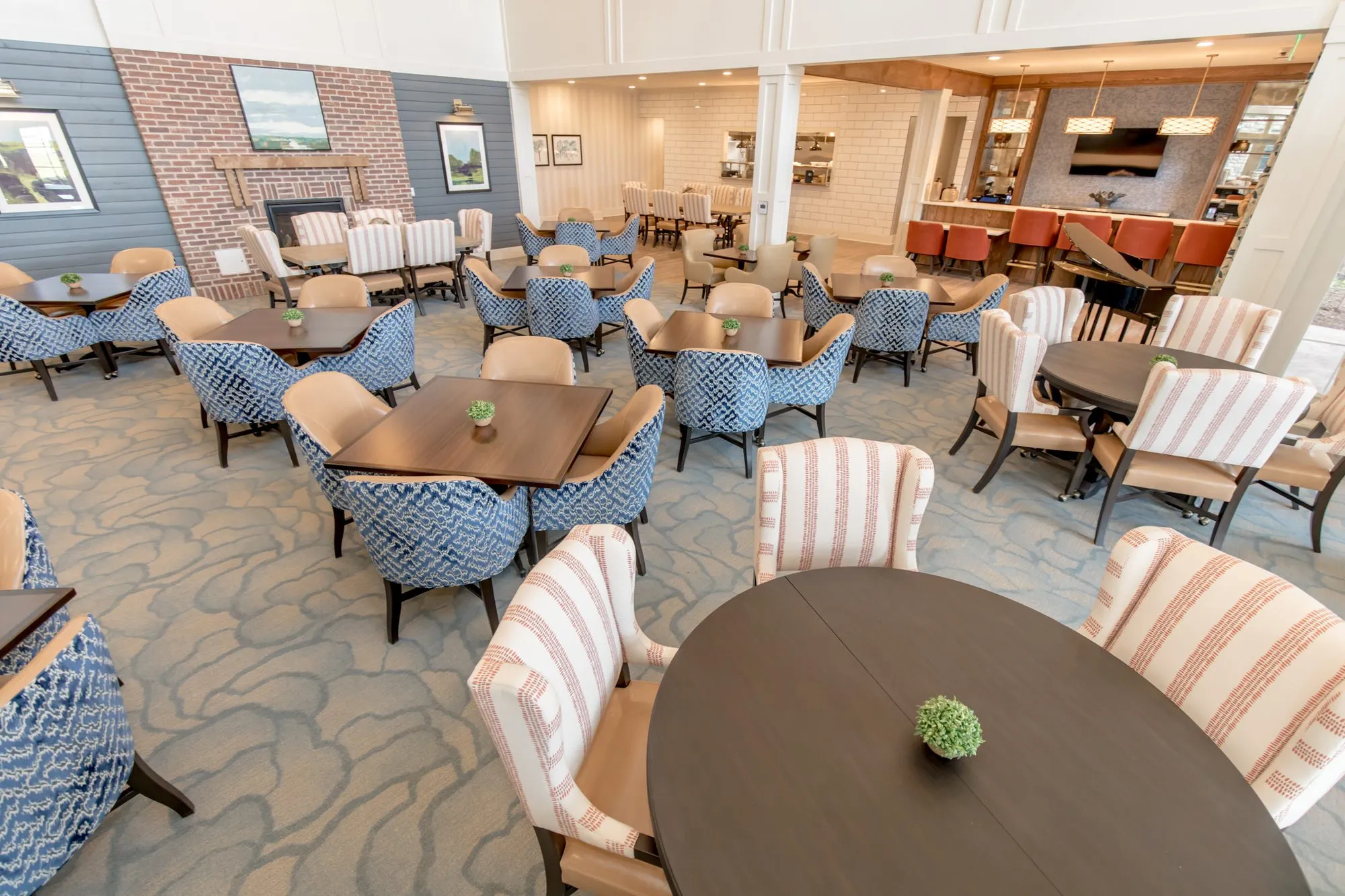 Senior living dining room with patterned and striped chairs at dark tables on a swirl carpet, brick fireplace and bar beyond