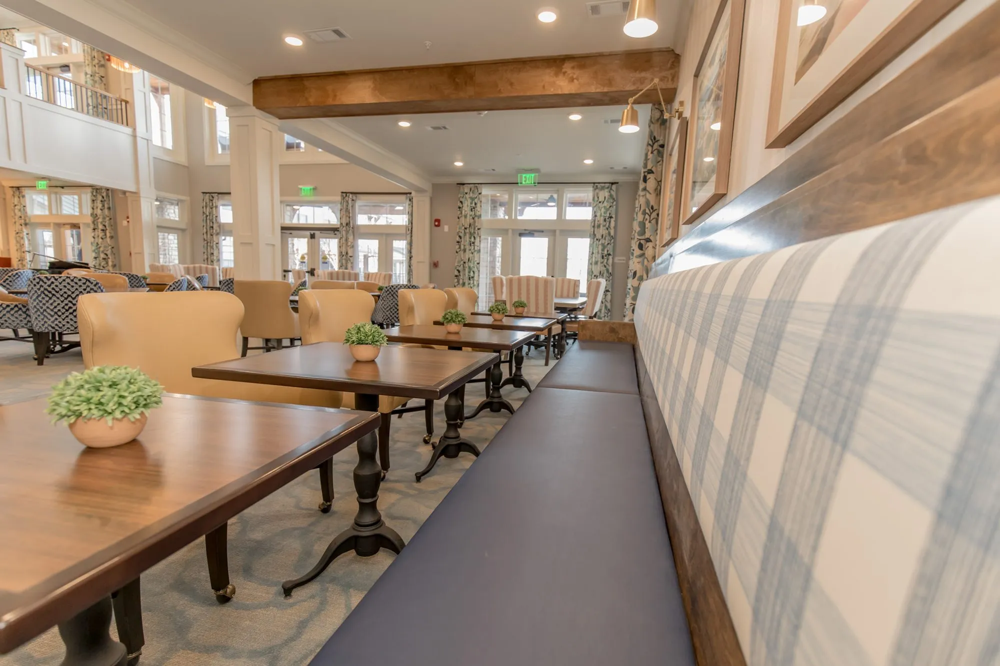Senior living dining room: long plaid banquette with a navy seat along dark wood tables, the two-storey room beyond