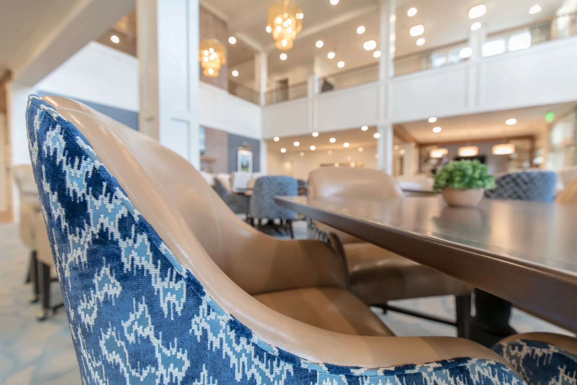 Senior living dining room detail: blue ikat and tan leather chair, crystal chandeliers in the two-storey volume beyond