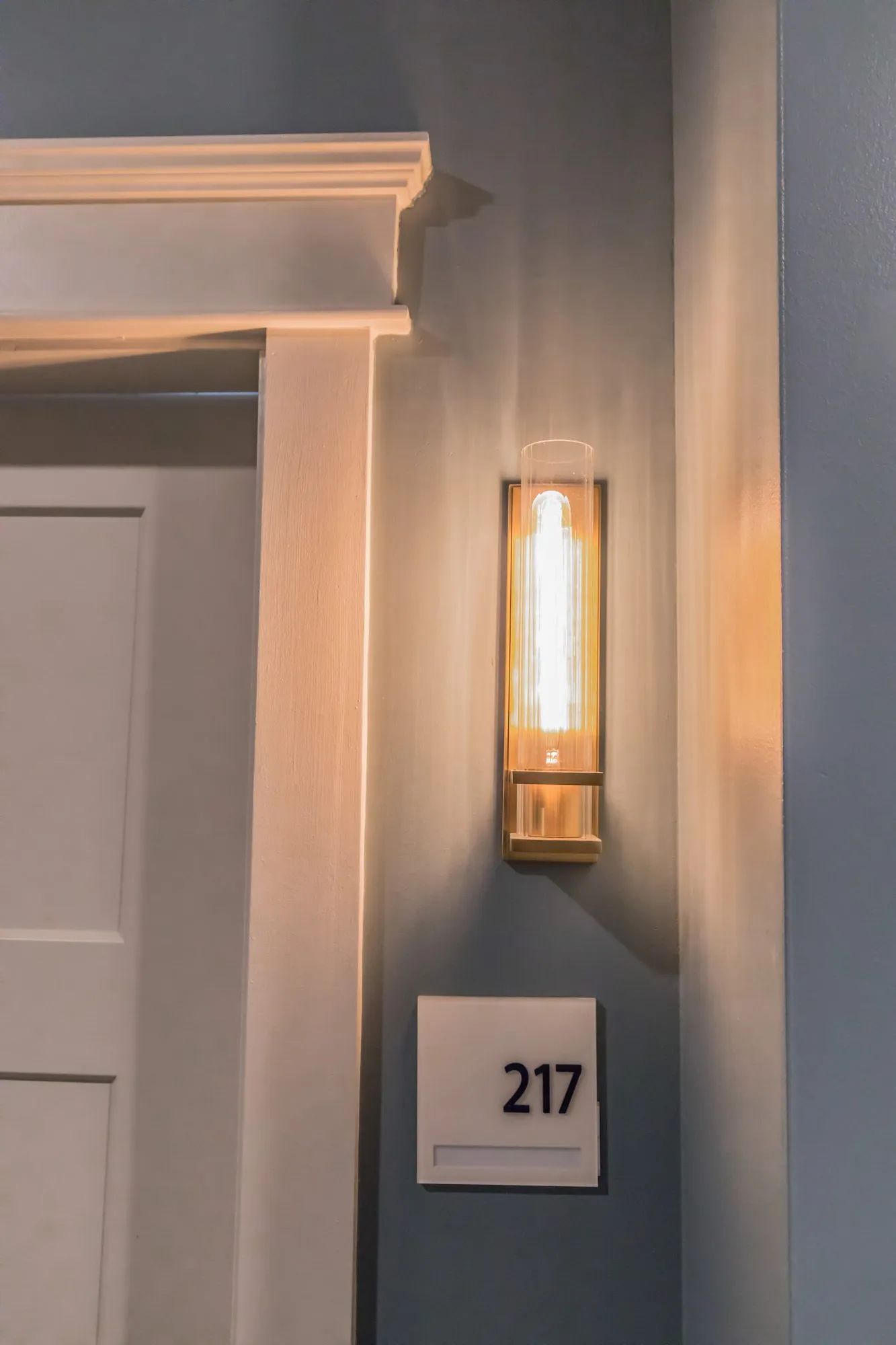 Senior living interior detail: brass and glass tube sconce beside white door trim and an apartment number plaque