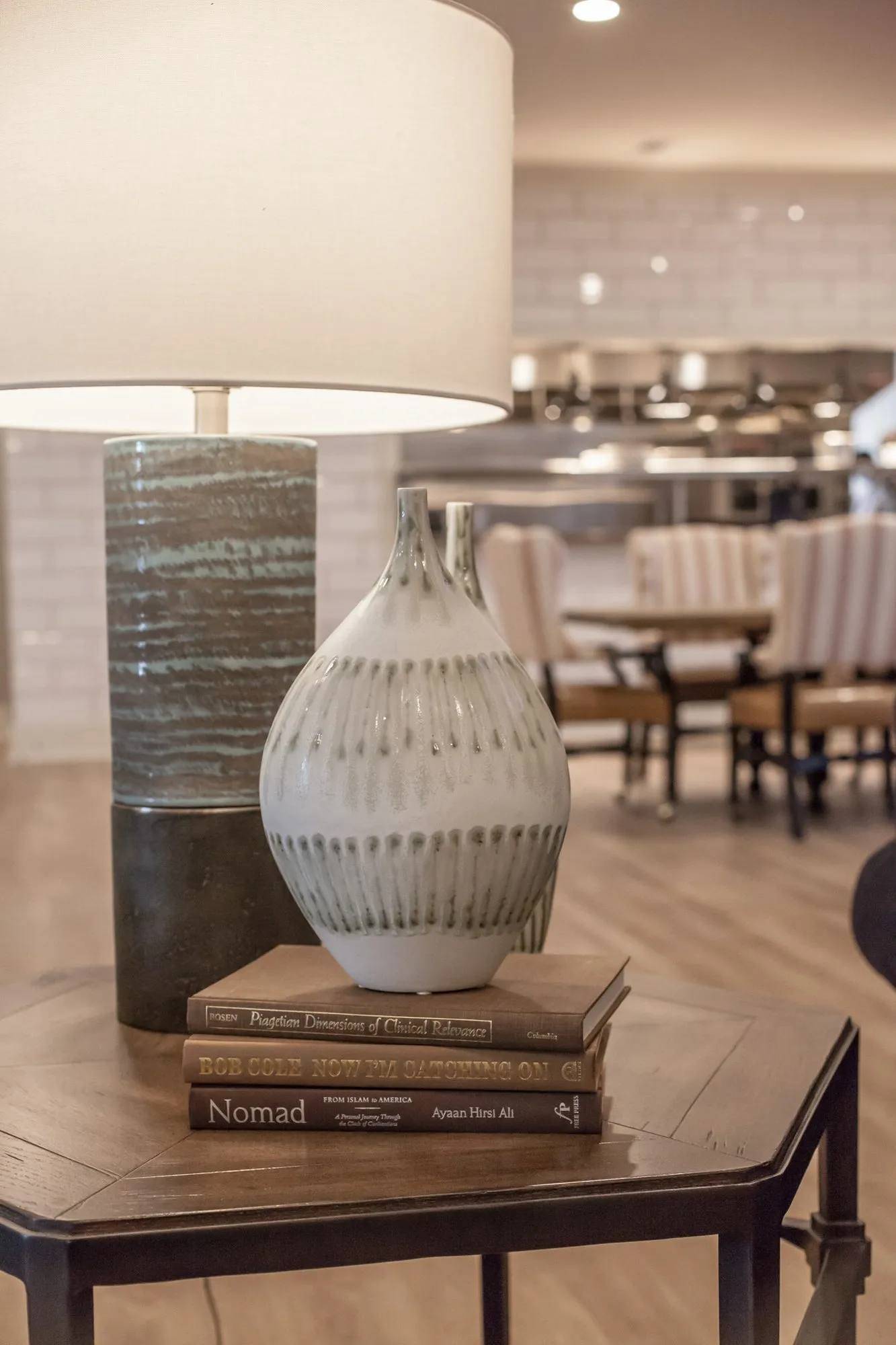 Senior living interior detail: striped vase and books on a hexagonal side table under a ceramic lamp, dining chairs beyond