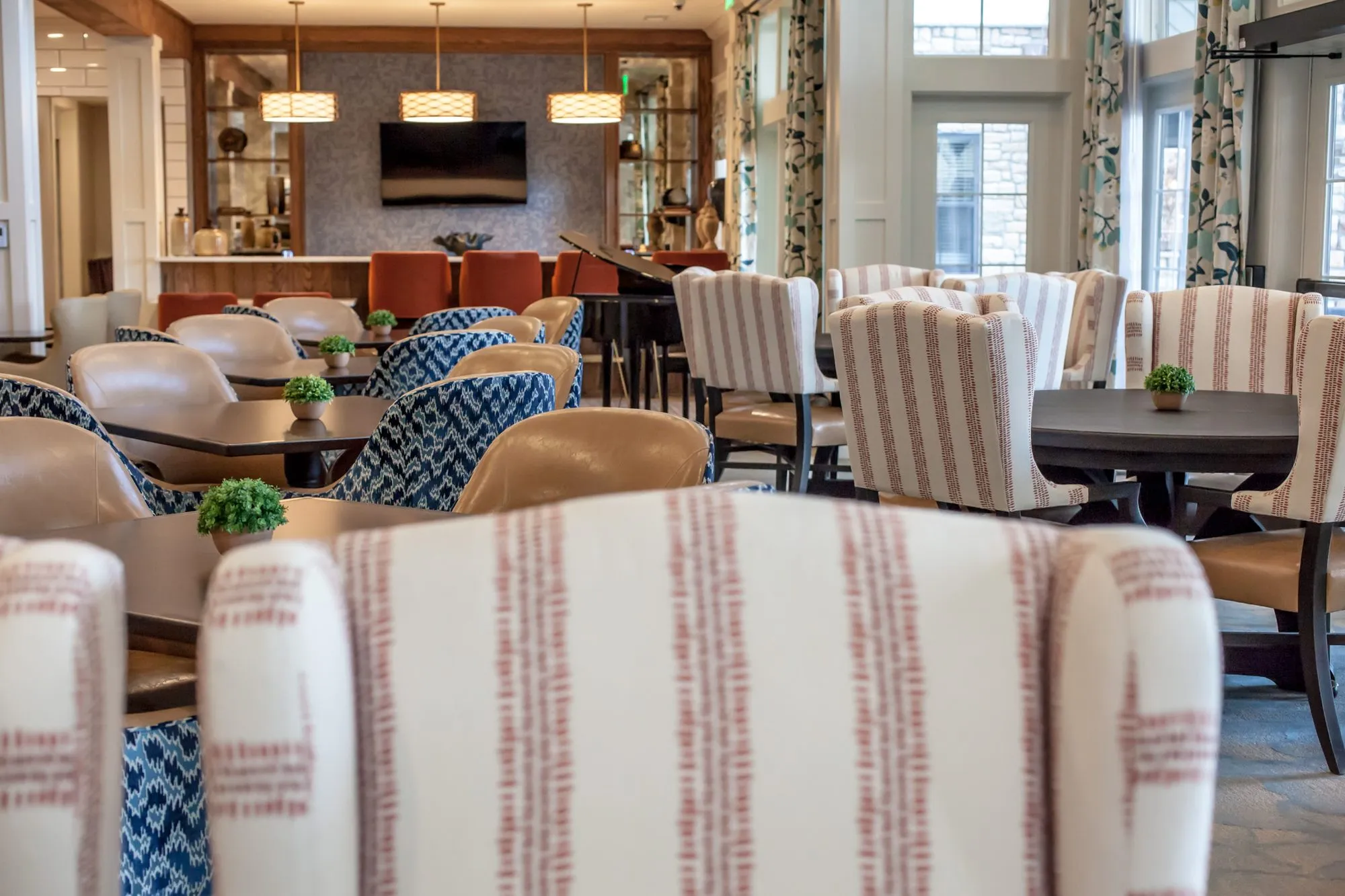 Senior living dining room with striped and blue patterned chairs at round tables, a bar with orange stools and piano beyond