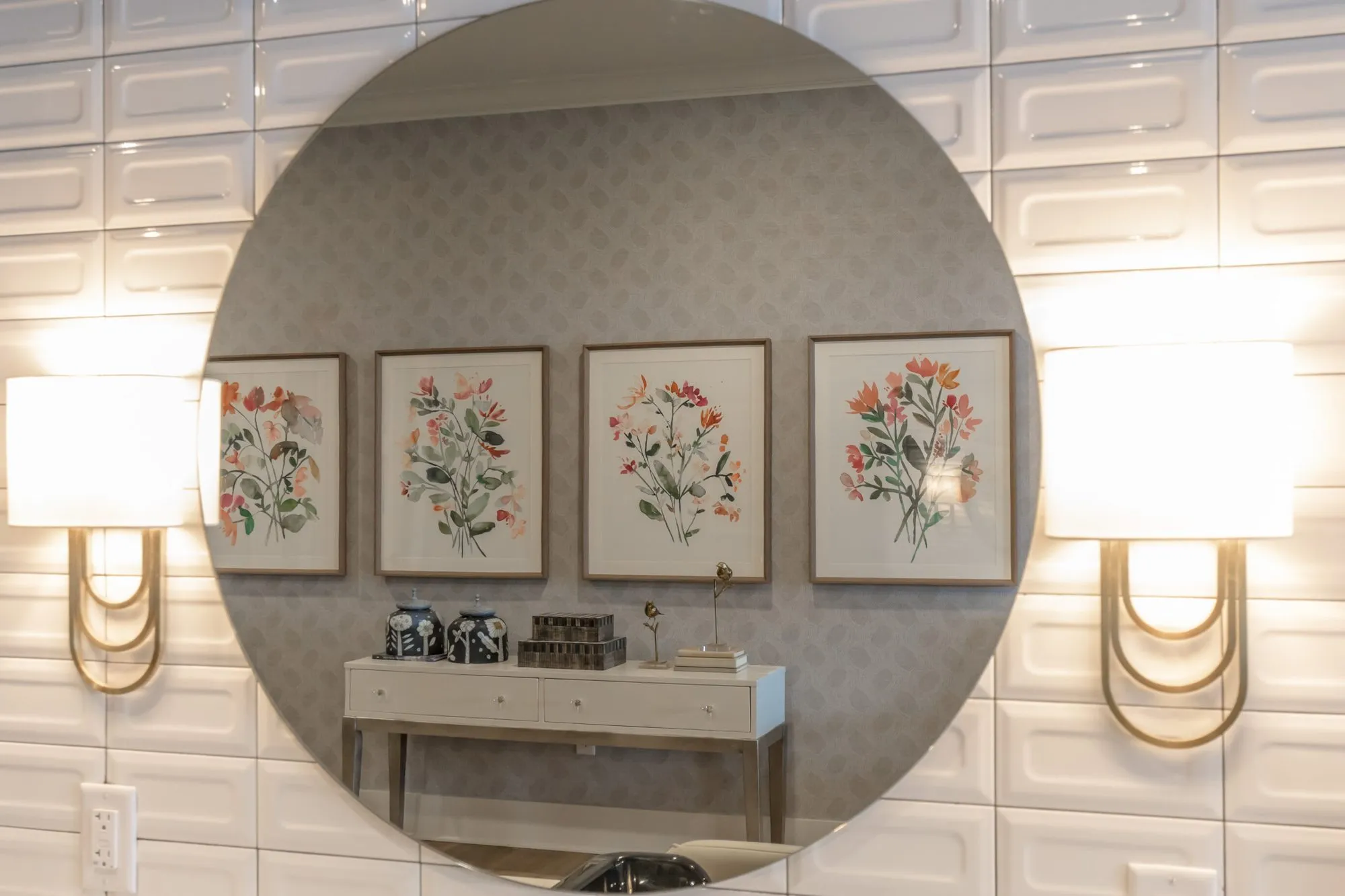 Senior living interior detail: round mirror between brass loop sconces on white tile, reflecting botanical prints