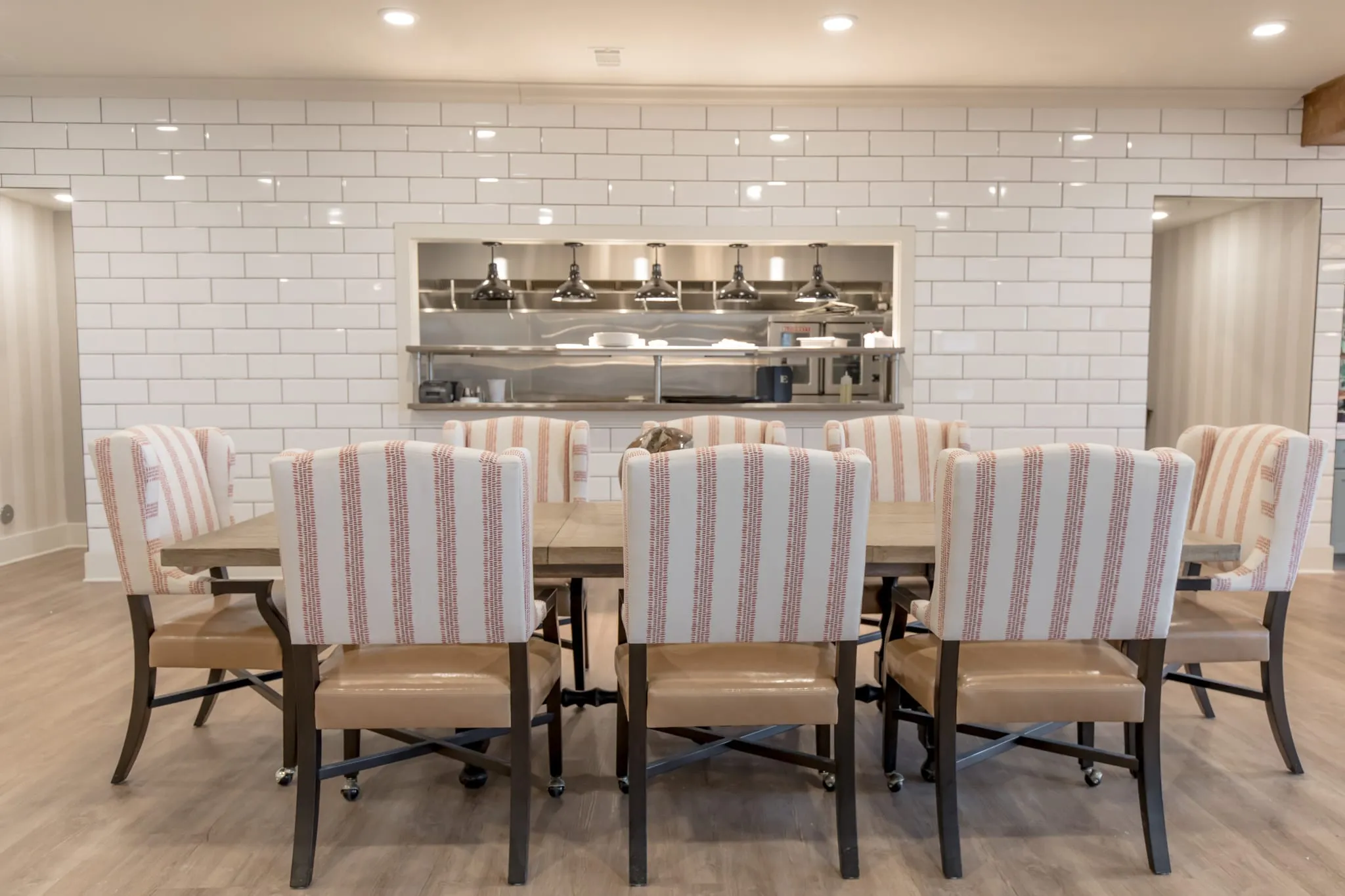 Senior living private dining room, red striped chairs at a long table before a subway tile wall with a kitchen pass-through