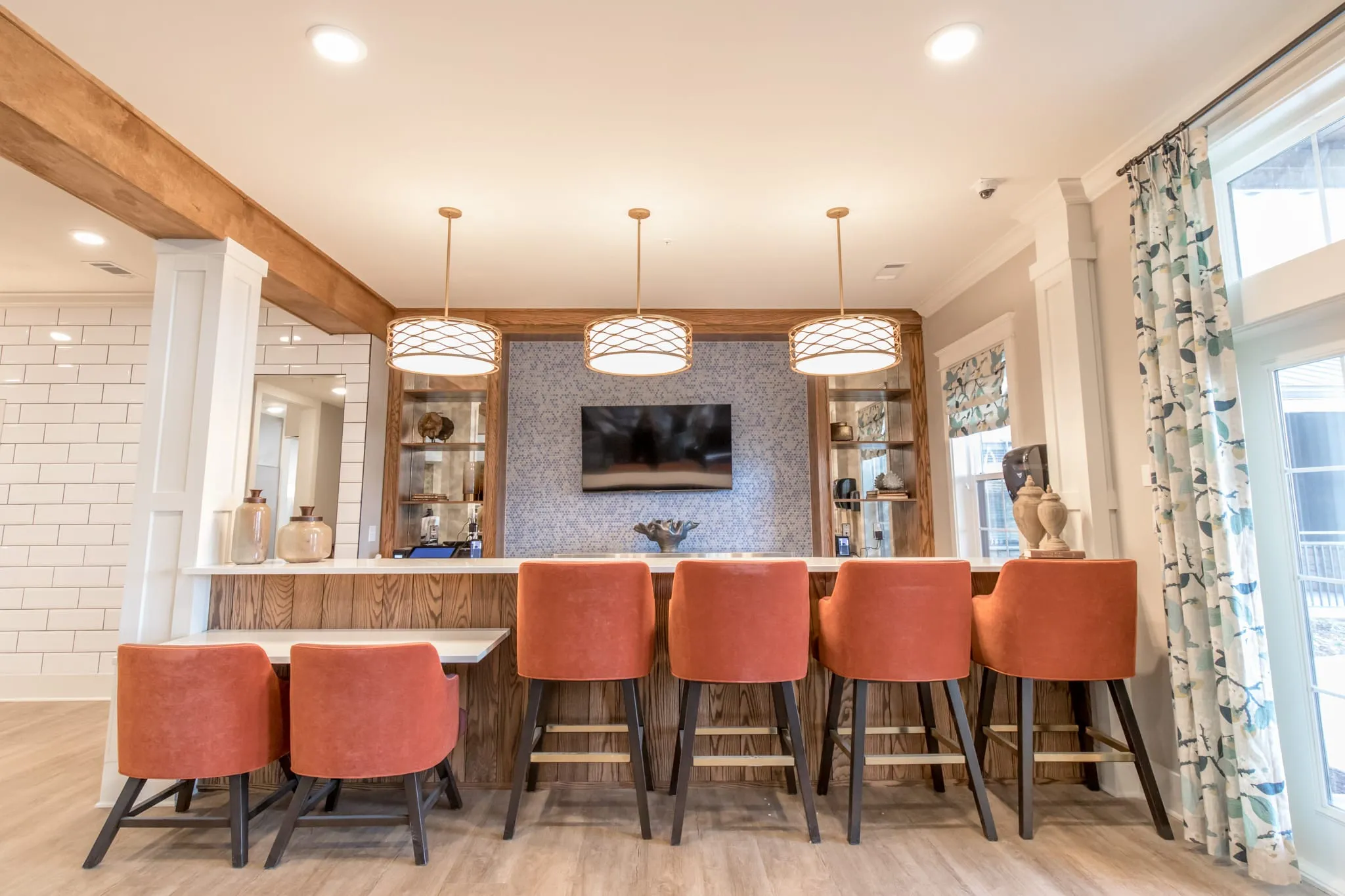 Senior living pub bar with coral leather stools along a wood bar under drum pendants and a timber beam