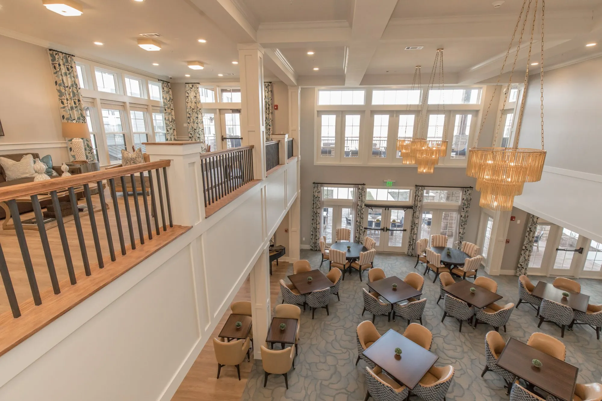 Senior living two-story great room seen from the mezzanine, amber glass chandeliers above the lounge and cafe seating