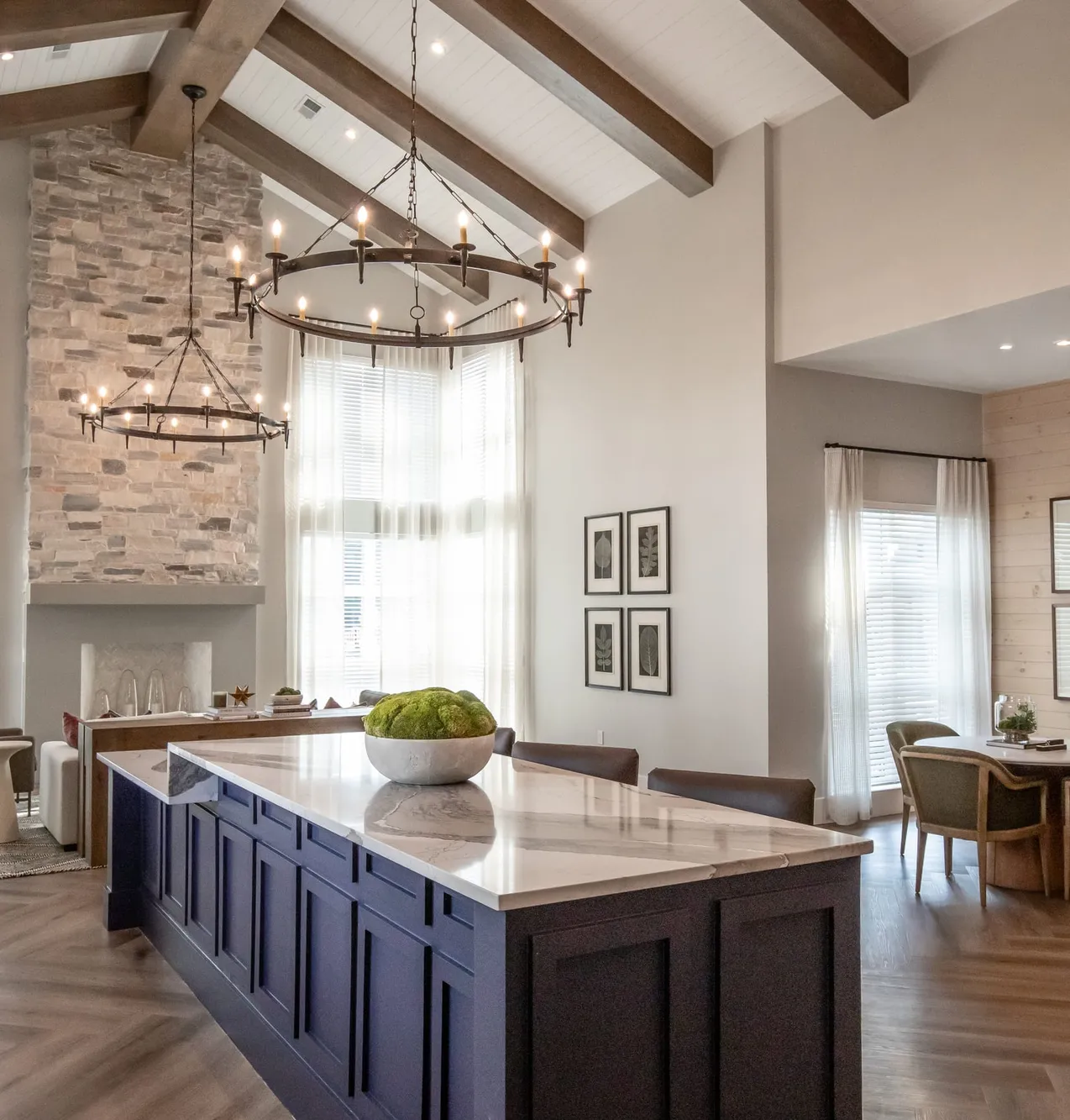 Clubhouse kitchen with a navy island under an iron ring chandelier, a stacked stone fireplace and a beamed ceiling