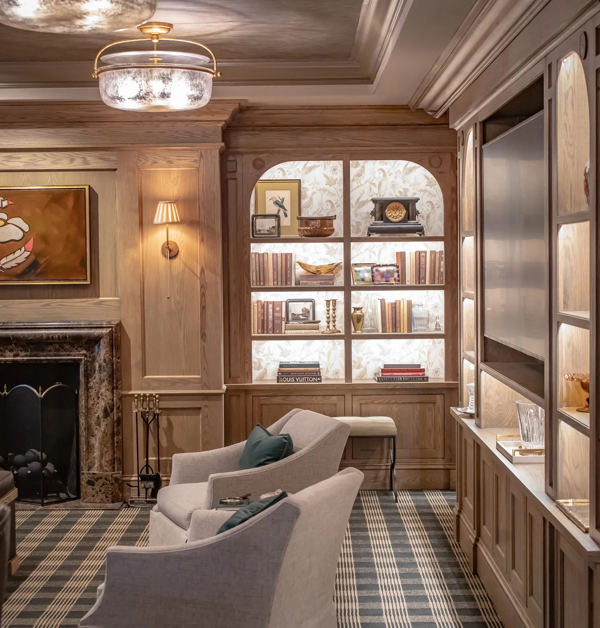Wood panelled club room with lit display shelving either side of a marble fireplace and cream armchairs on a checked rug