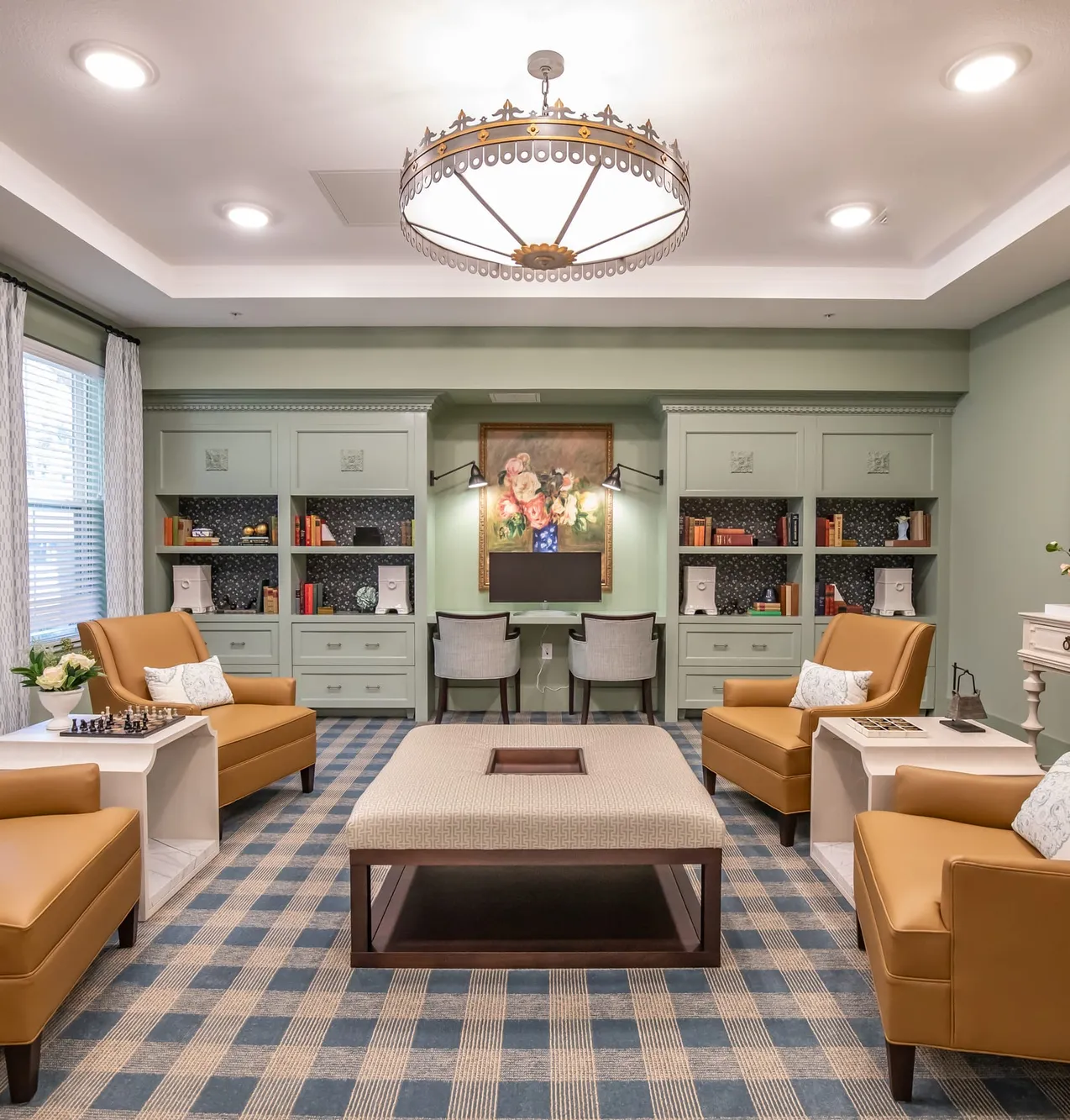 Library with sage green built-in shelving, orange wingback chairs on a blue checked rug and a drum pendant overhead