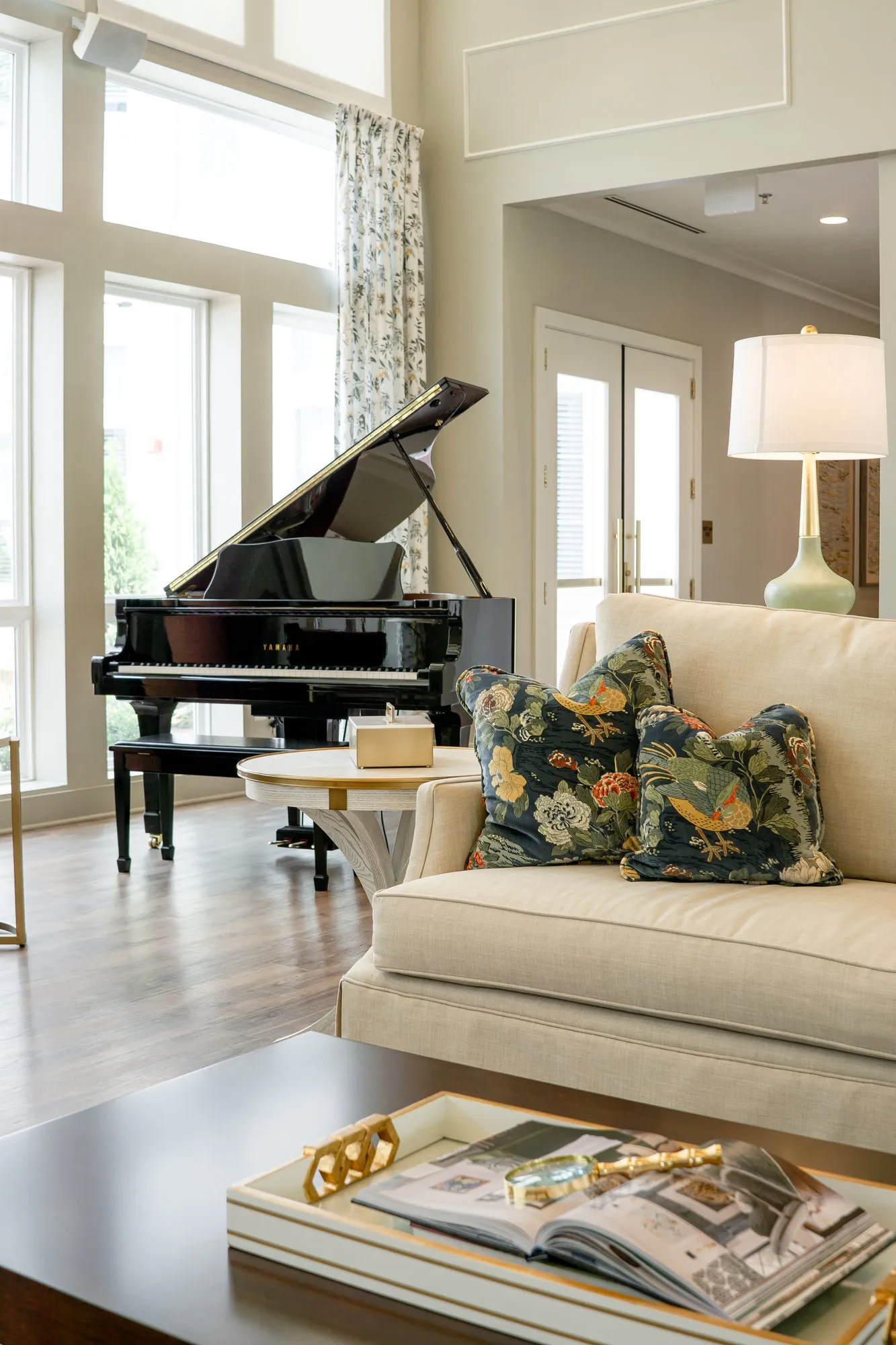 Senior living great room with a cream sofa and navy bird print pillows beside a black grand piano at the tall windows