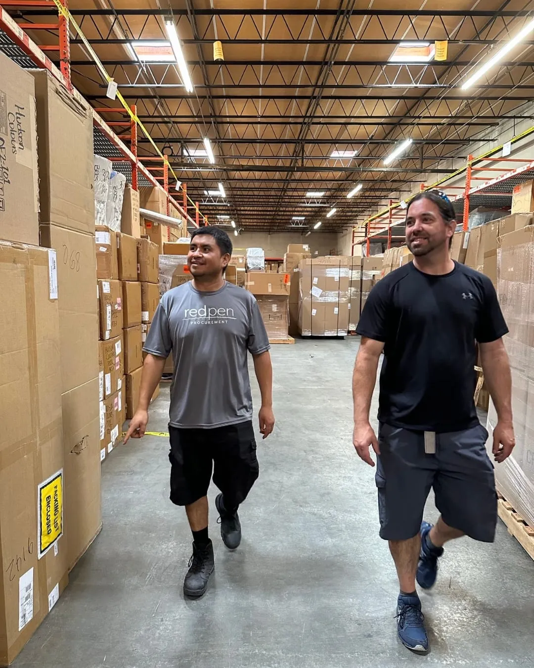 Two men walking down a warehouse aisle between pallets of stacked cardboard boxes under steel roof trusses