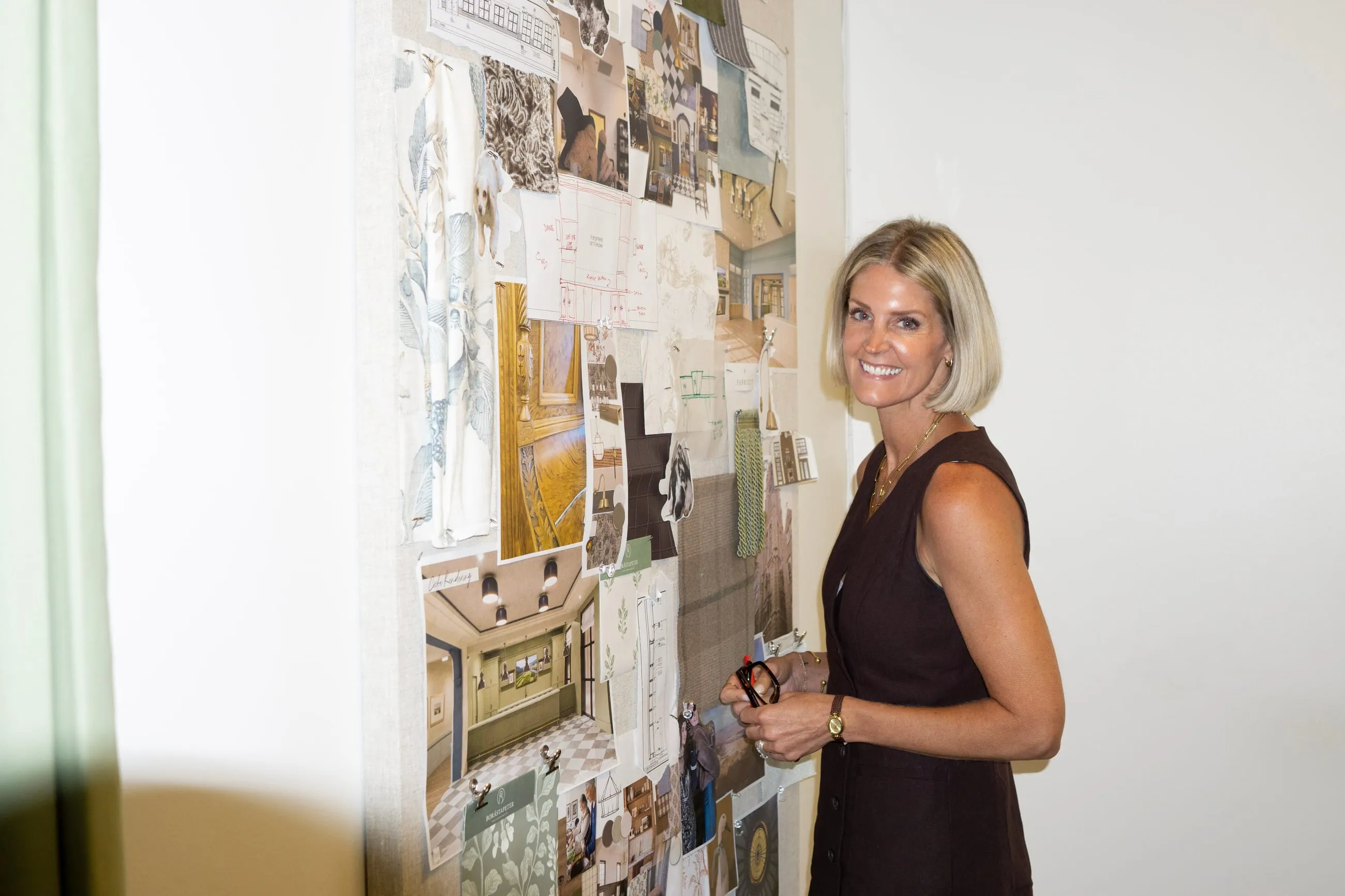 Melissa Banko in a black dress, standing beside a studio wall pinned with tear sheets, sketches and material samples