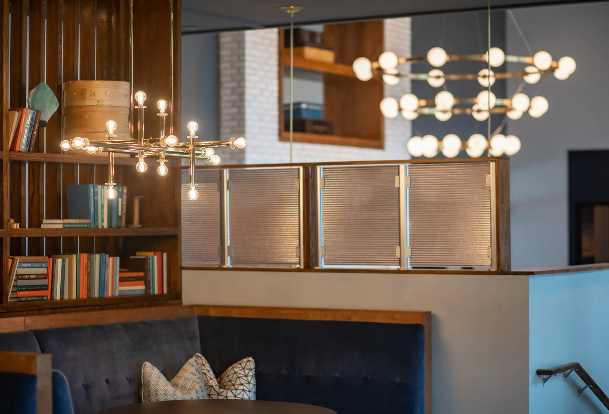 Multifamily amenity clubhouse lounge with brass globe chandeliers, walnut shelving full of books and a navy velvet banquette below pleated shades