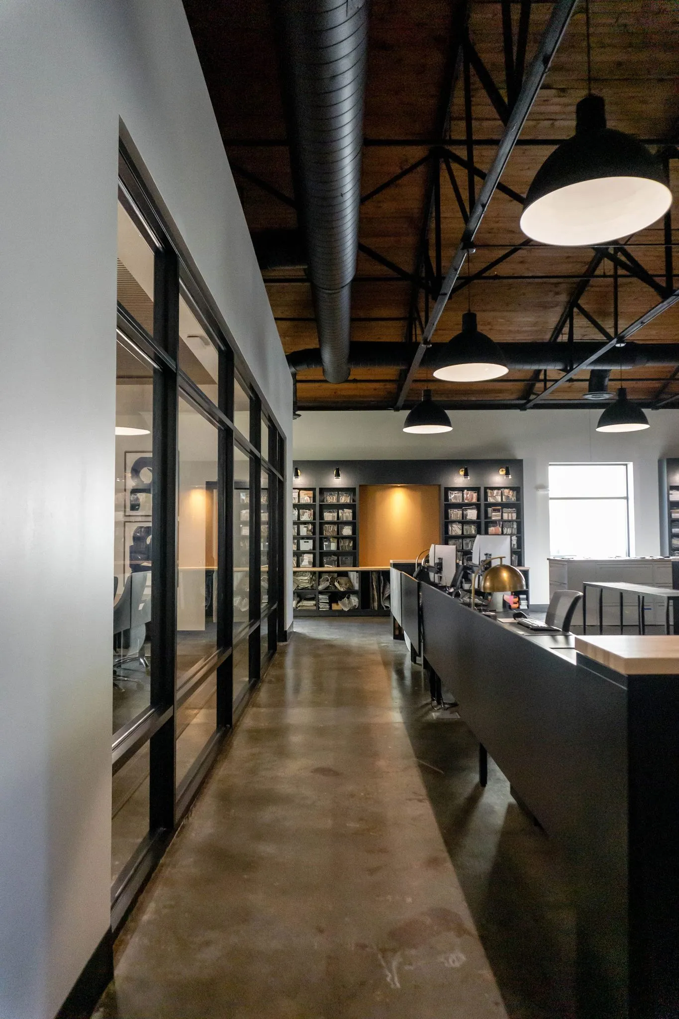Office corridor with black framed glazing along a polished concrete floor and dome pendants under the wood barrel ceiling