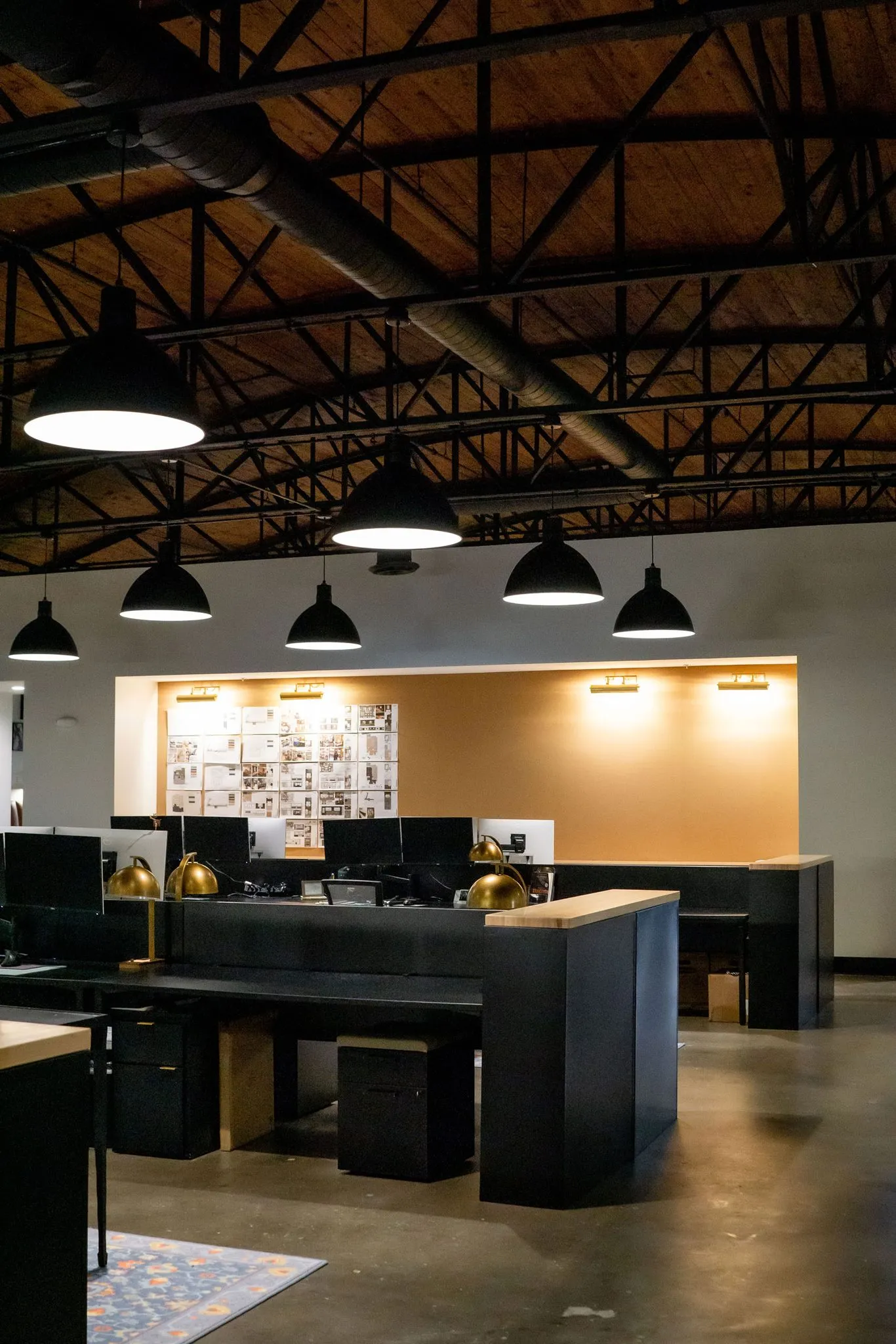 Office open studio of black desks with monitors and brass lamps under dome pendants and steel trusses, pinned boards beyond