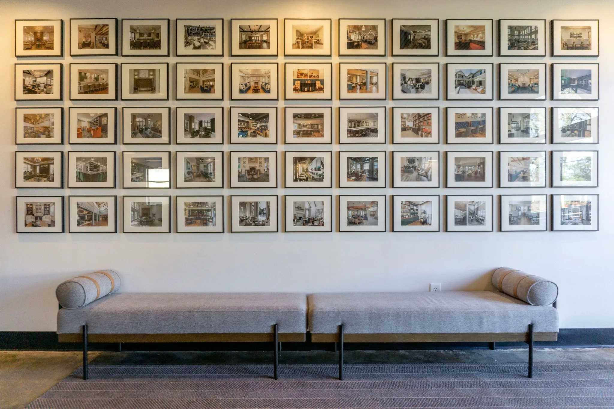 Office interior detail: grid of about fifty framed project photographs above a long grey upholstered bench with bolsters