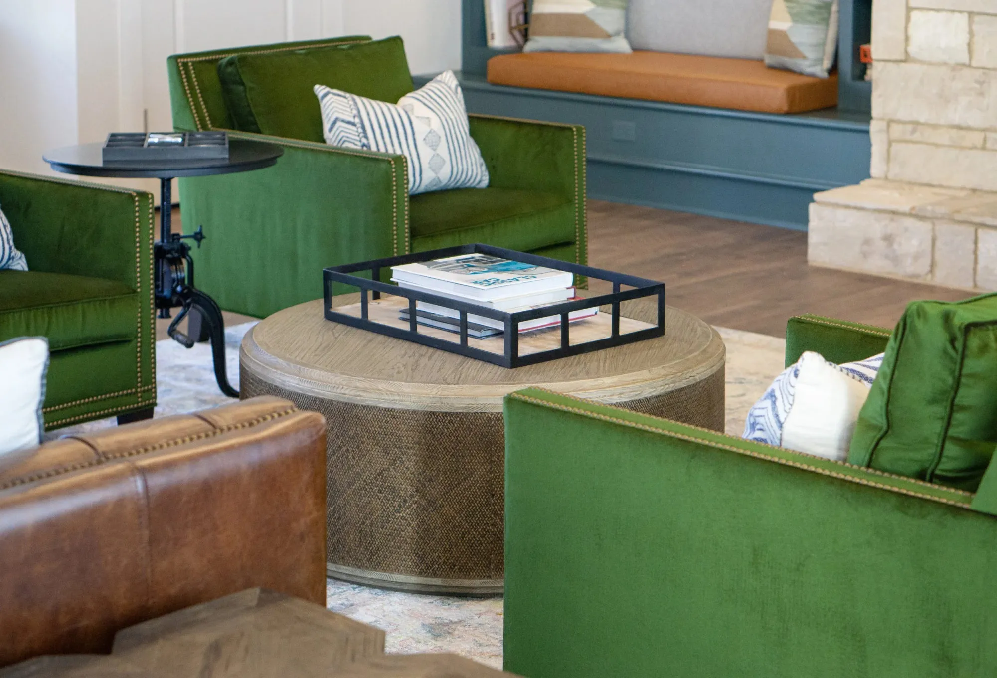 Multifamily amenity fireside seating with emerald velvet armchairs around a rattan-wrapped table, a cognac leather sofa and stone fireplace