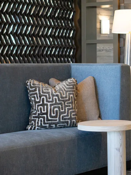 Grey banquette sofa with a black and white cut velvet pillow against a dark sculptural wall panel by a pale wood side table