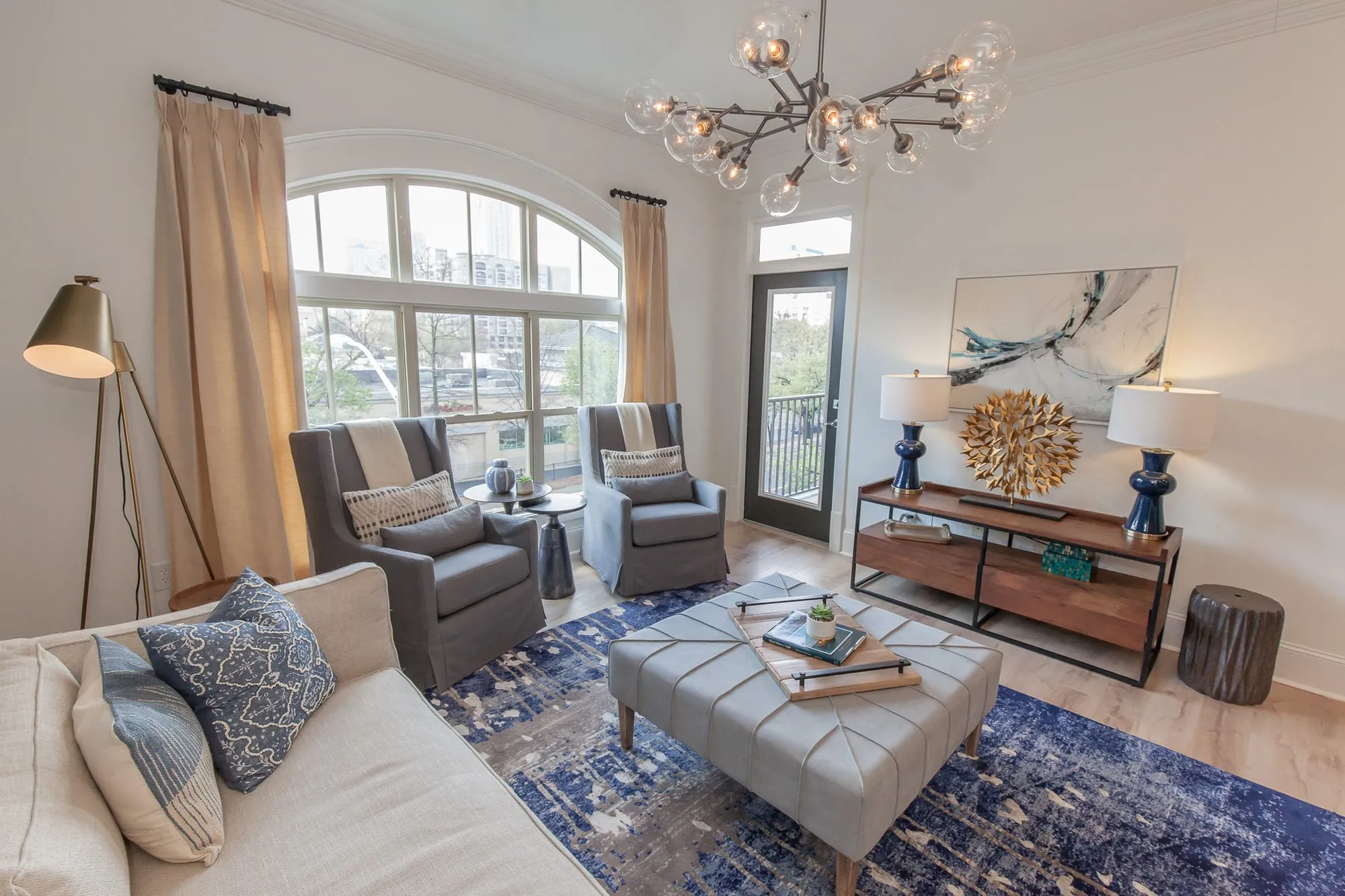Multifamily apartment living room from behind a cream sofa, grey swivel chairs by the arched window under a globe chandelier