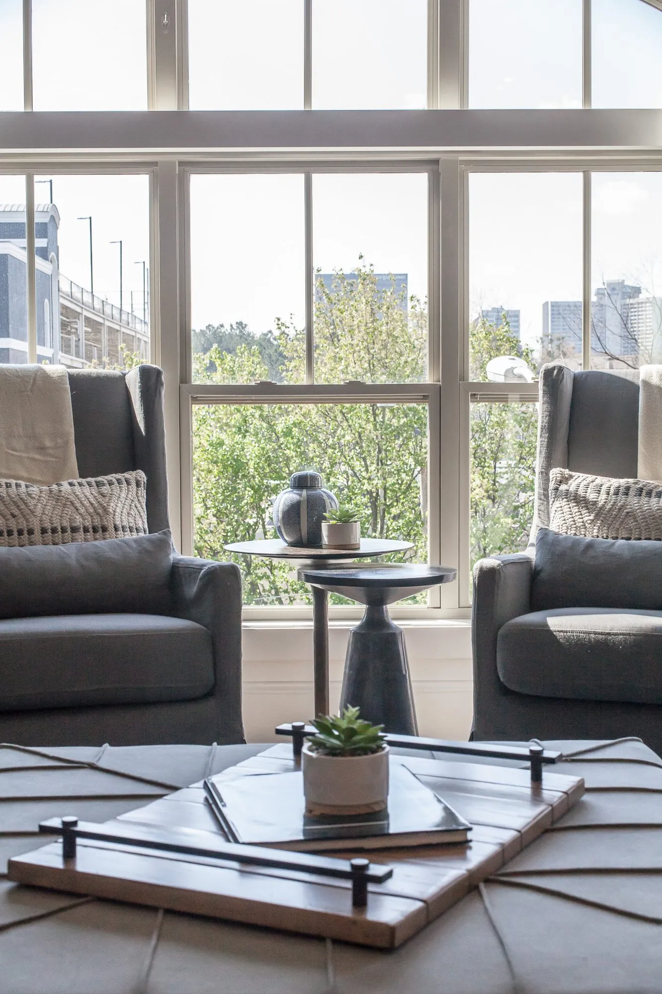 Multifamily interior detail: two grey wingback chairs and a pedestal table before tall windows looking over trees to the city