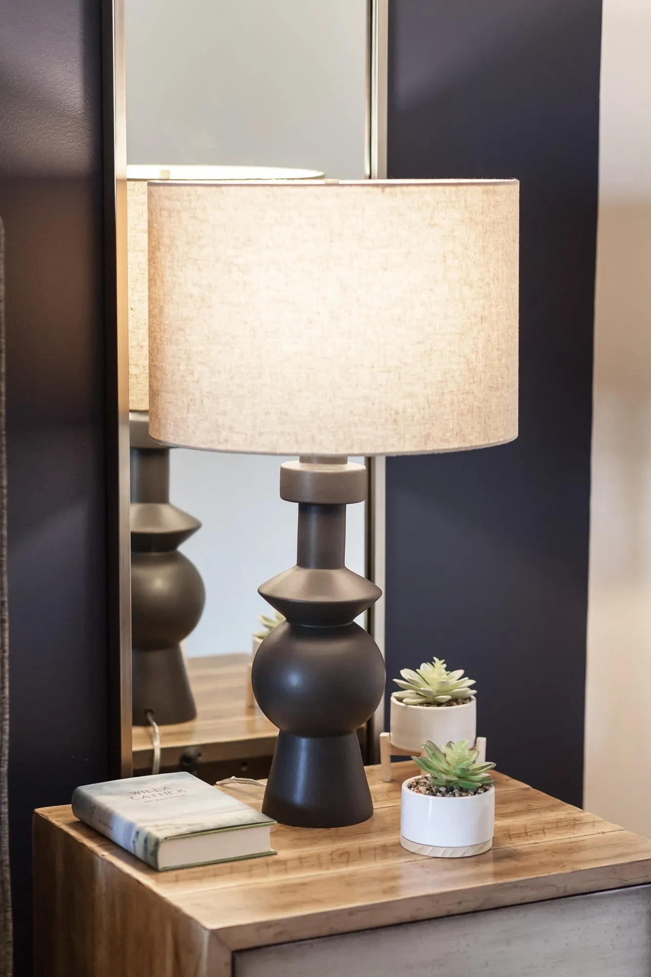Multifamily apartment bedside detail: black turned lamp with a linen shade, succulents and a book against a navy wall