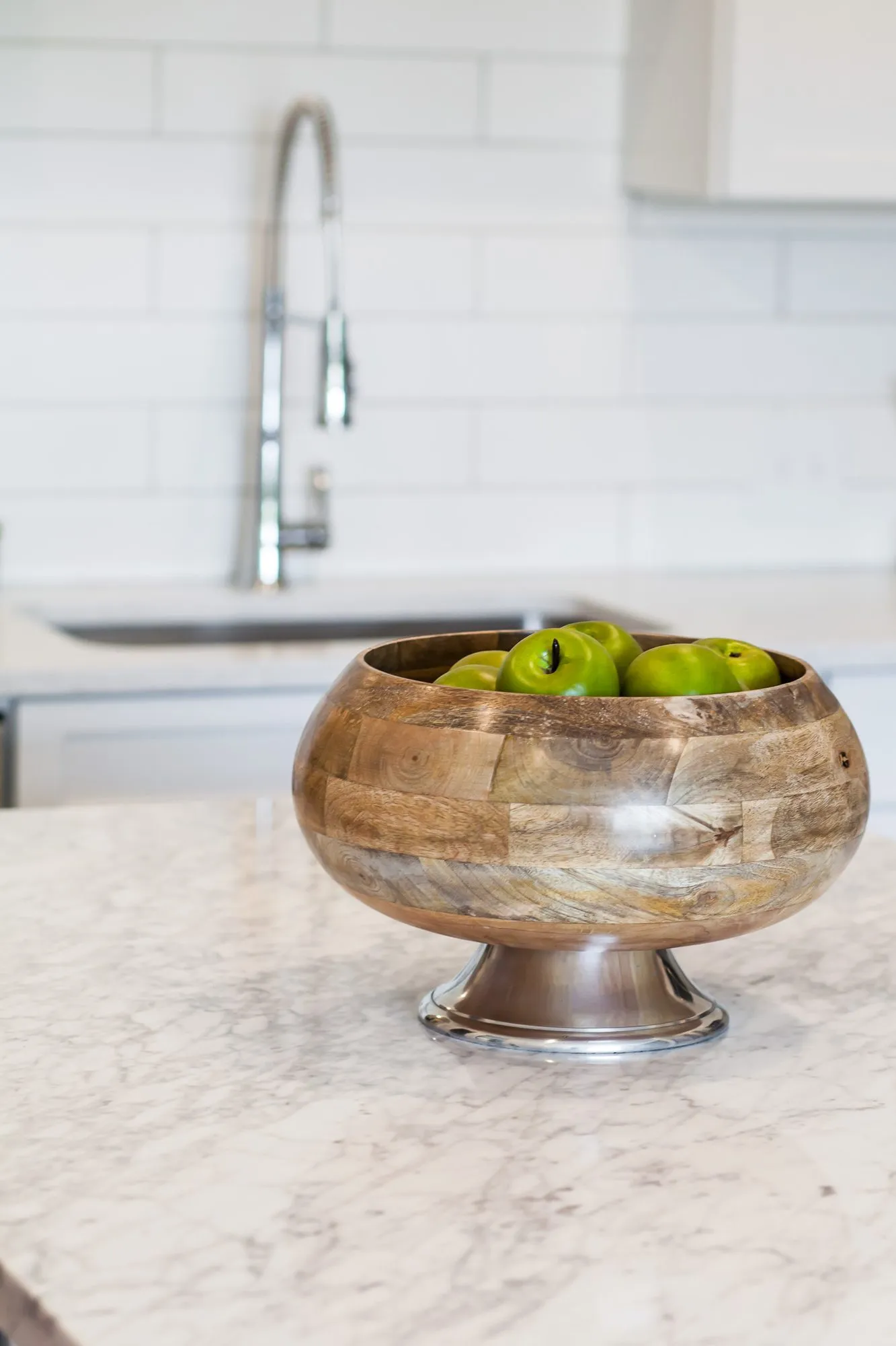 Multifamily kitchen detail: wooden footed bowl of green apples on a marble countertop, white subway tile and chrome faucet