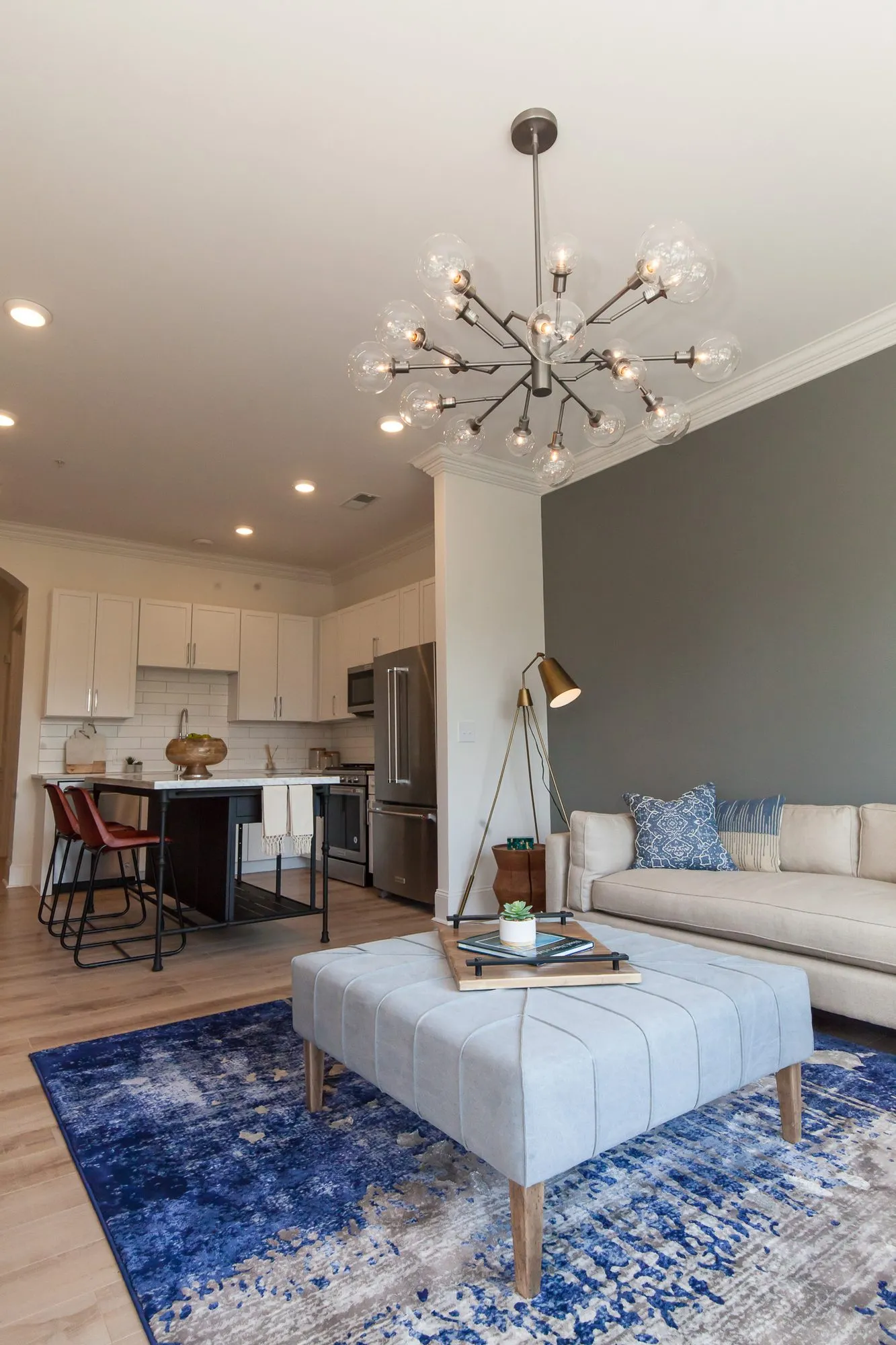 Multifamily apartment open plan living area with a glass globe chandelier above a tufted ottoman, kitchen behind