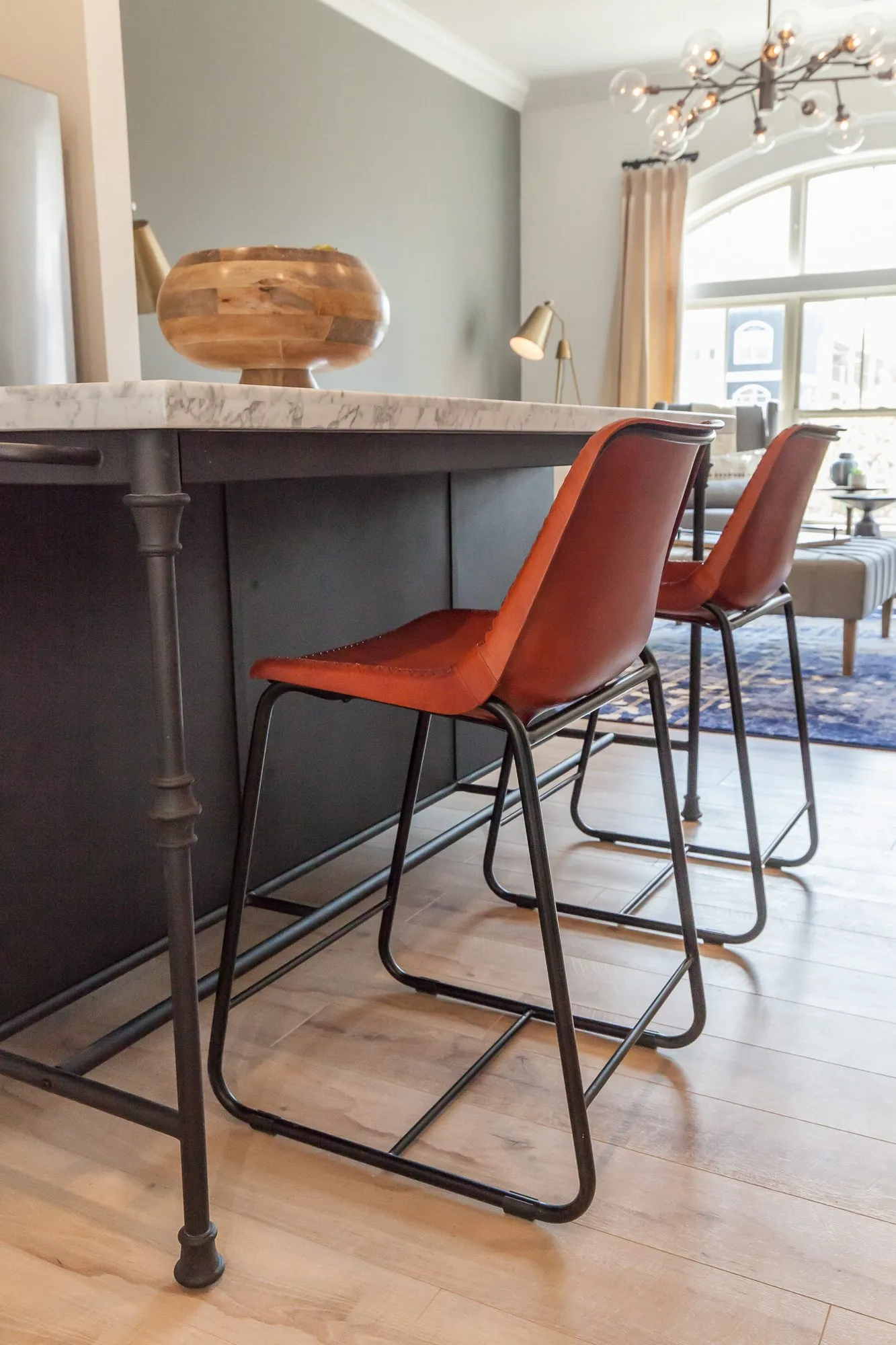 Multifamily apartment kitchen island with rust leather barstools on an iron base, the living room and arched window beyond