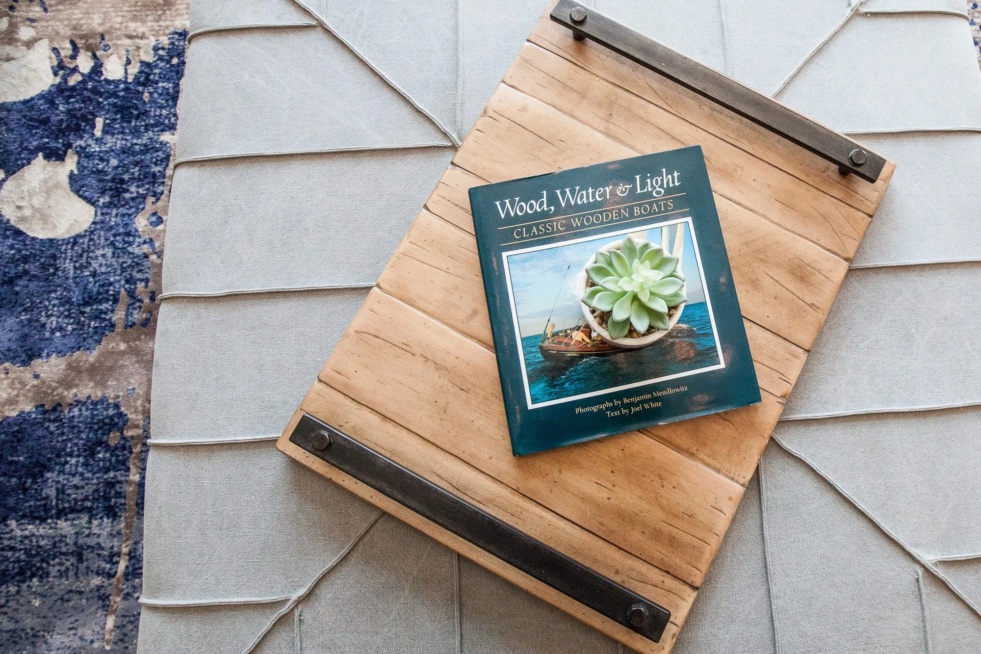 Multifamily interior detail: overhead view of a reclaimed wood tray with iron handles holding a book on a grey tufted ottoman