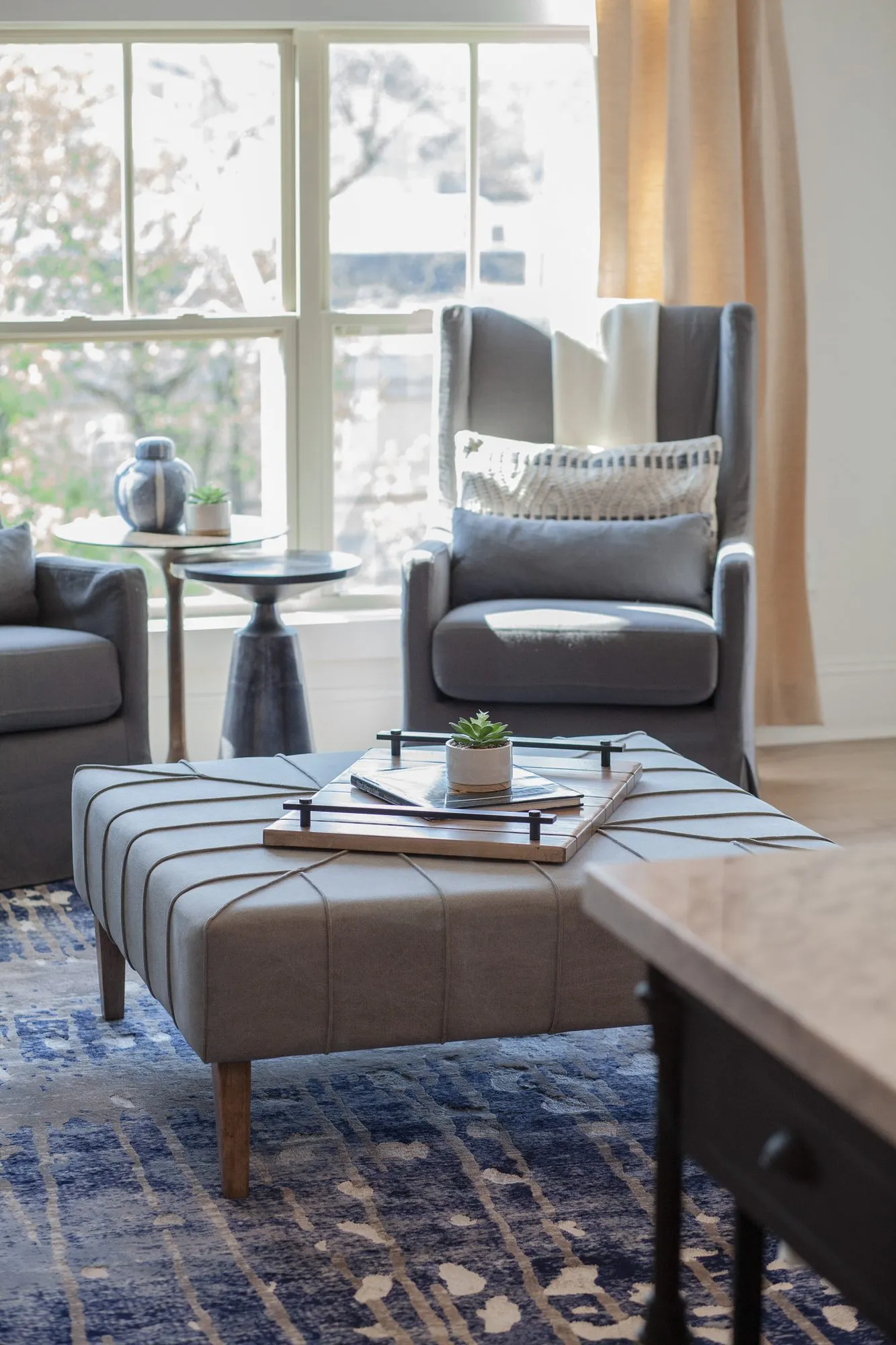 Multifamily apartment living room with a grey tufted ottoman on a blue rug, wingback chairs by the window