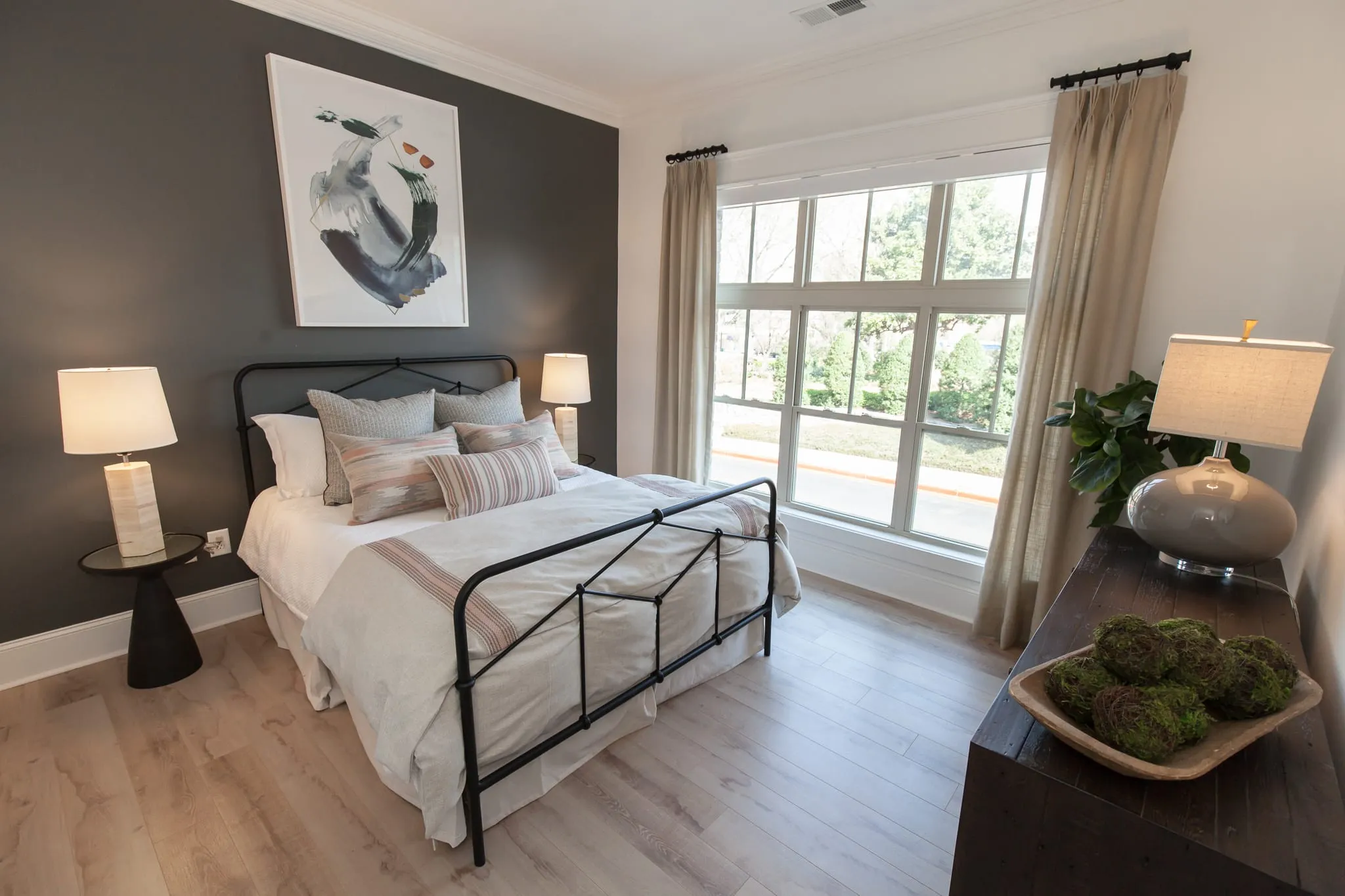 Multifamily apartment bedroom with a black iron bed against a charcoal accent wall beside tall windows with a leafy view
