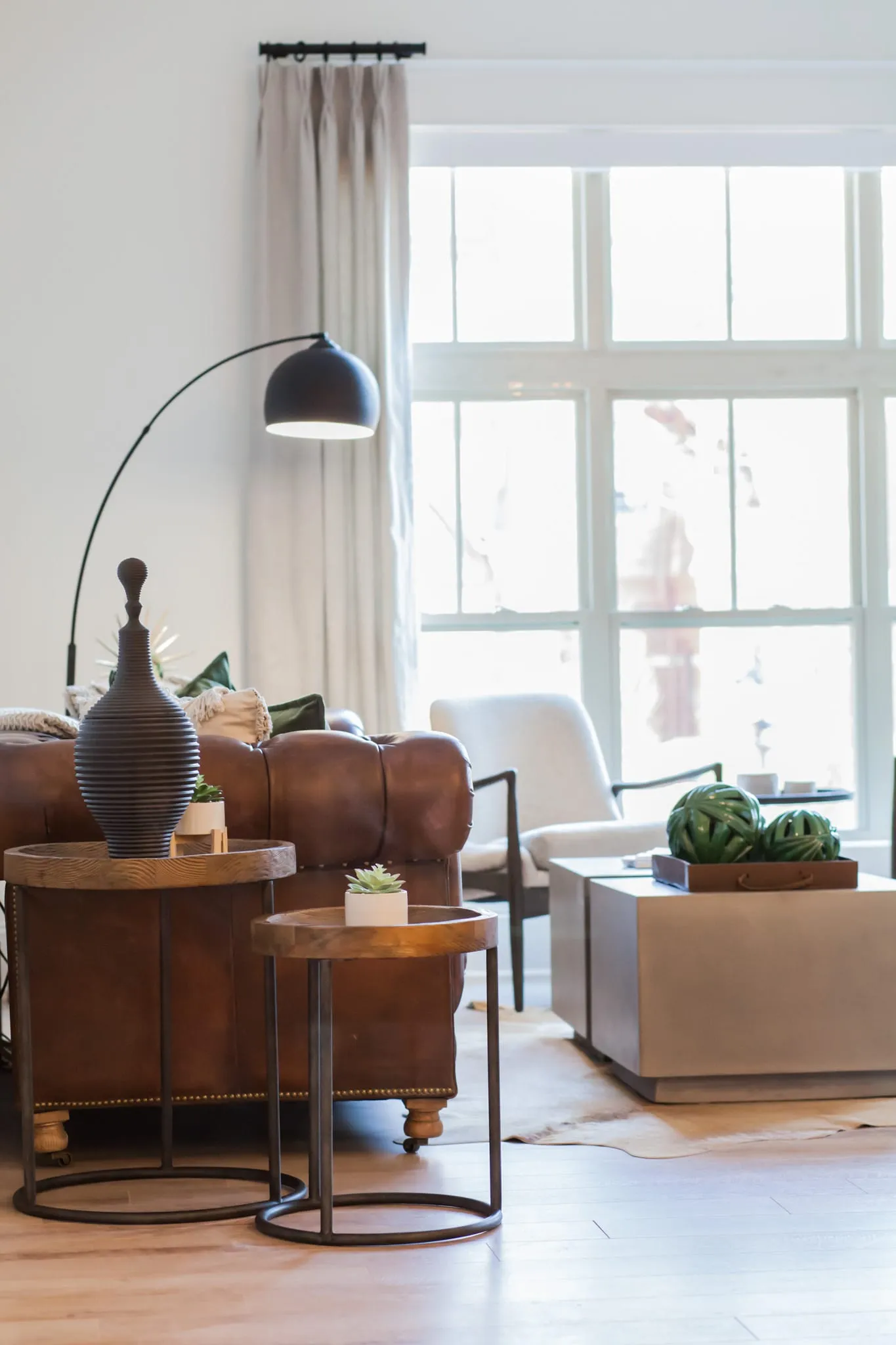 Multifamily apartment living room with a brown leather sofa, round nesting tables and an arc lamp beside a tall window