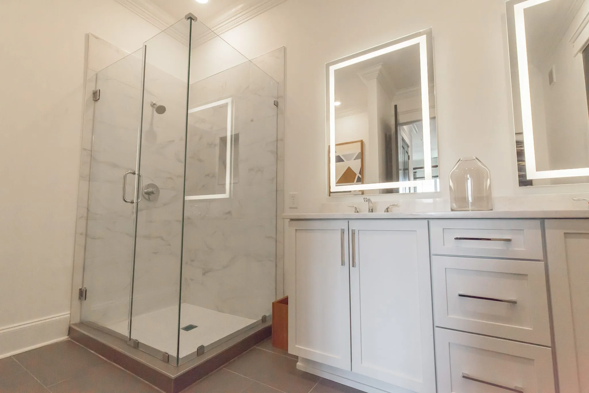 Multifamily apartment bathroom with a frameless glass corner shower in marble tile beside a backlit mirror