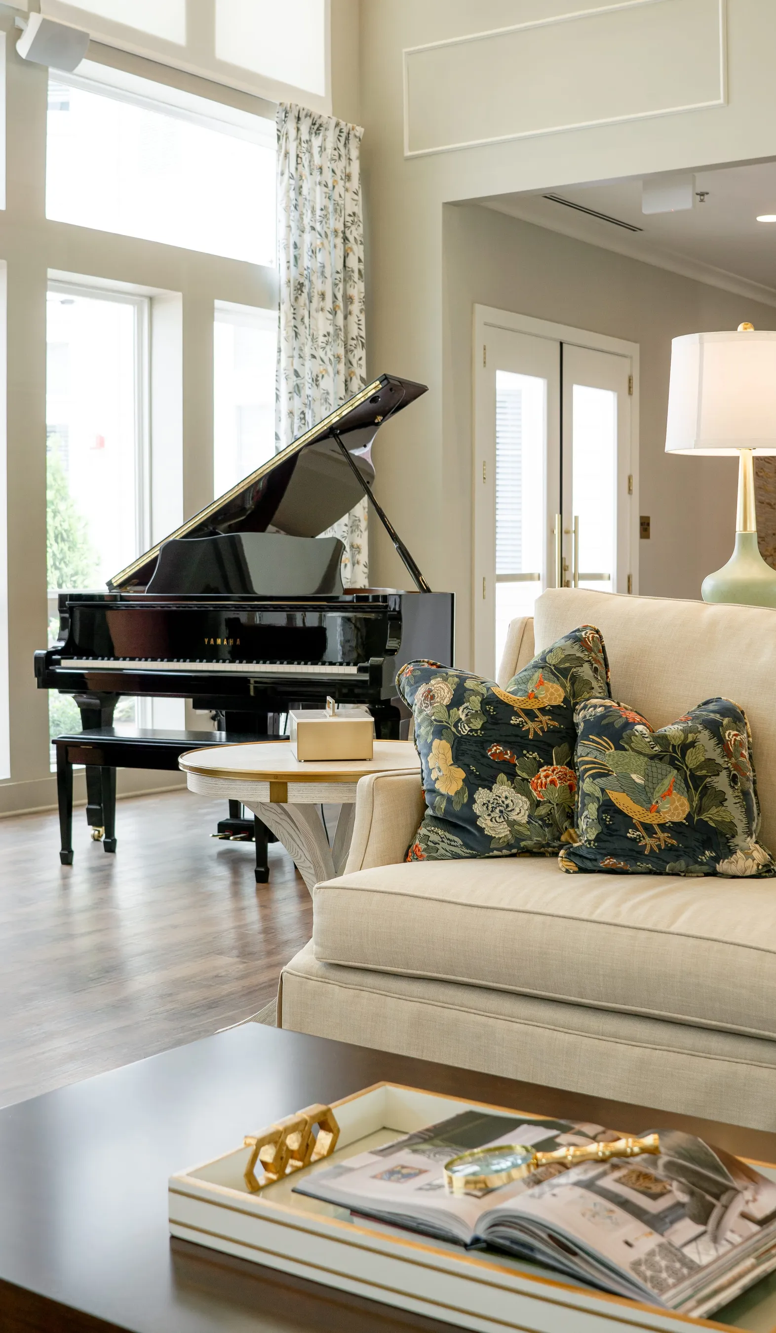 Senior living great room with a black grand piano at tall windows beside a cream sofa with navy bird print pillows