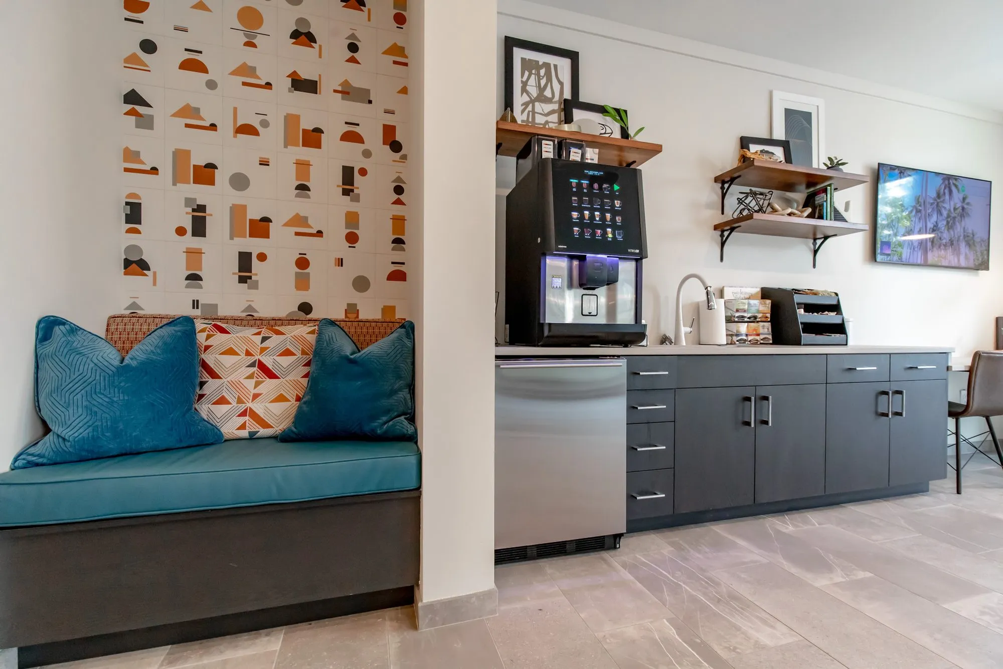 Multifamily amenity bench nook with teal cushions and geometric wallpaper beside a coffee station of charcoal cabinets