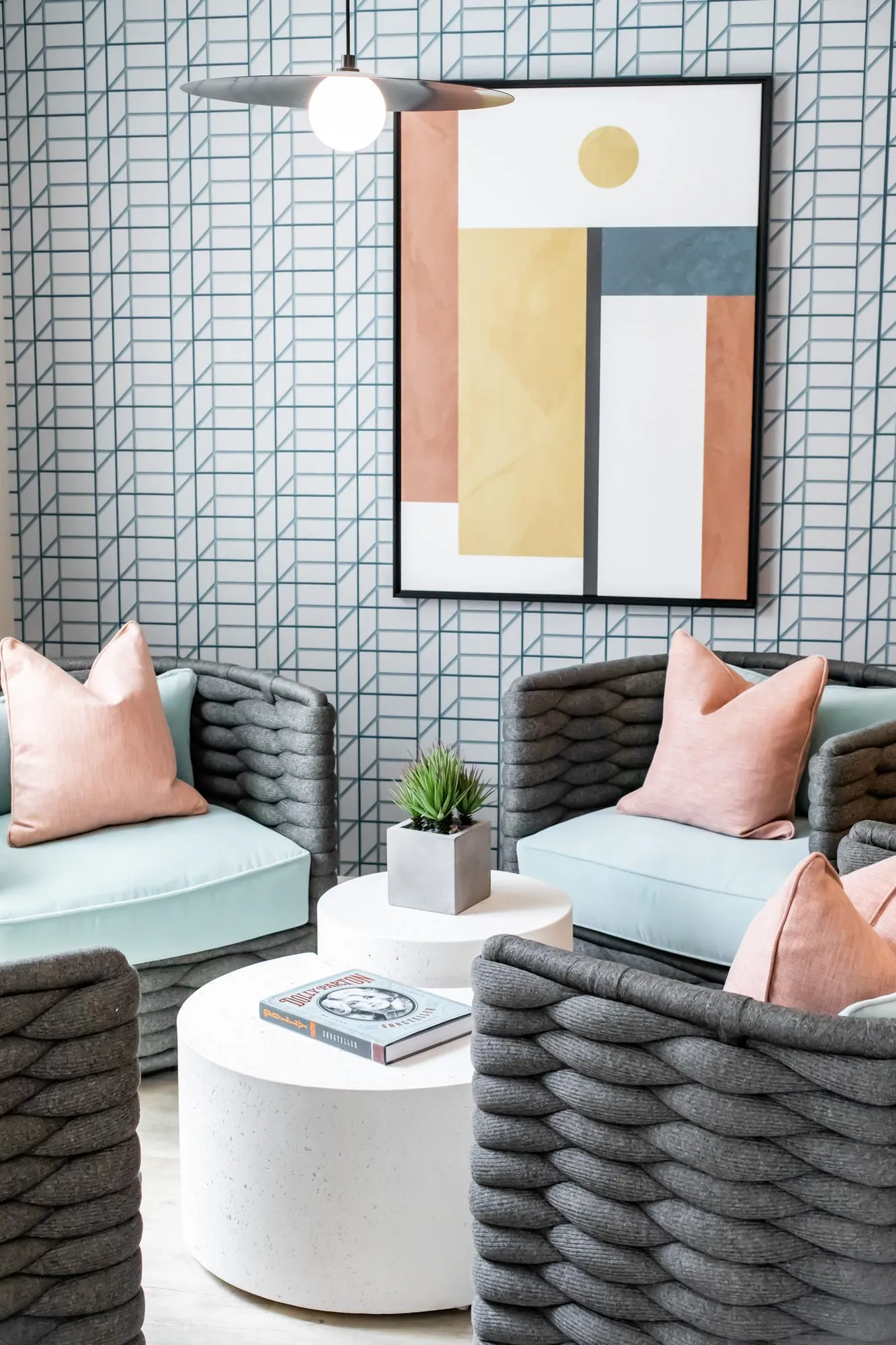 Multifamily amenity lounge with woven charcoal chairs and pink pillows around white drum tables, blue geometric wallpaper