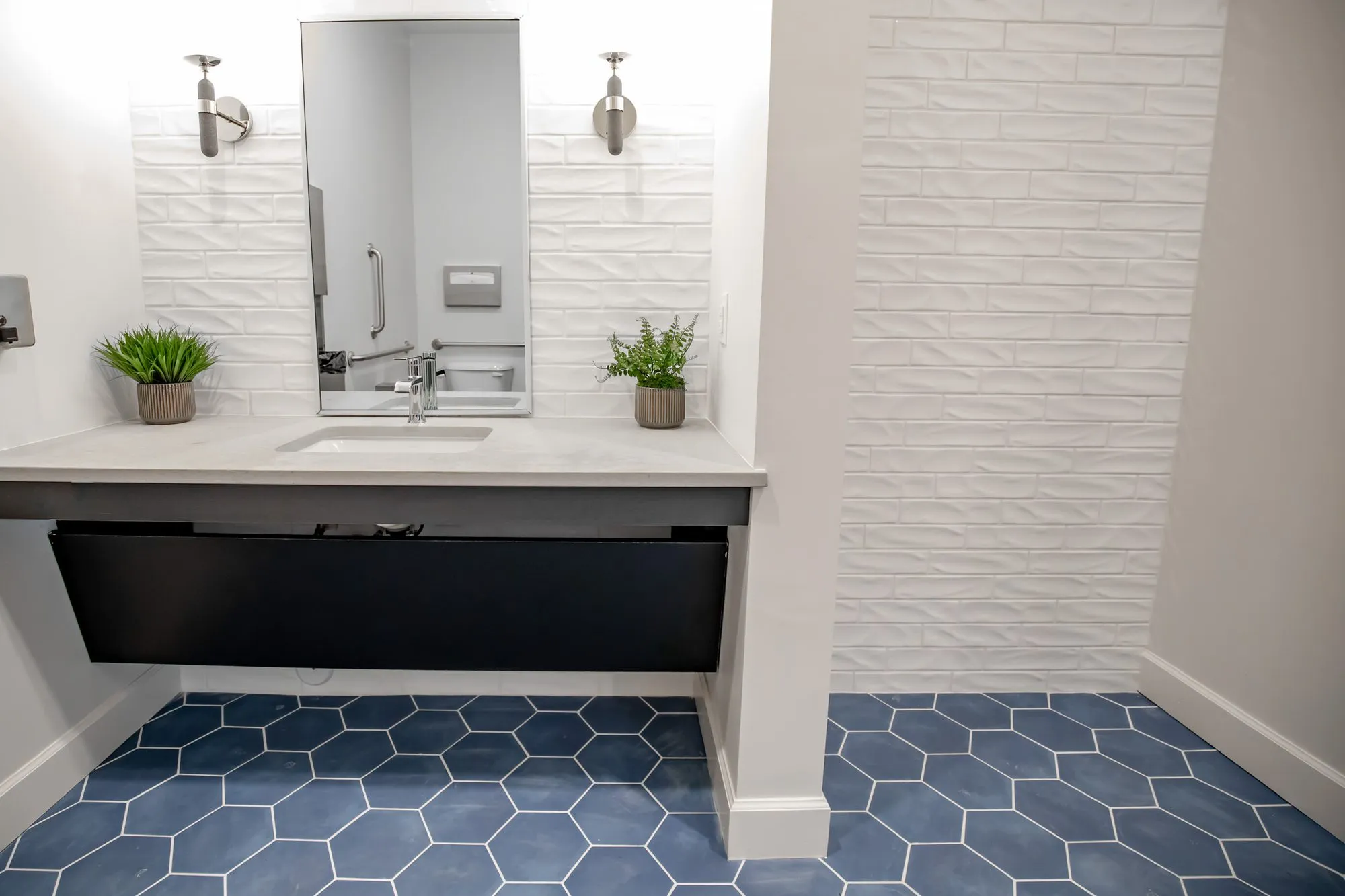 Multifamily amenity restroom with a floating stone vanity and rectangular mirror on white textured tile, navy hexagon floor
