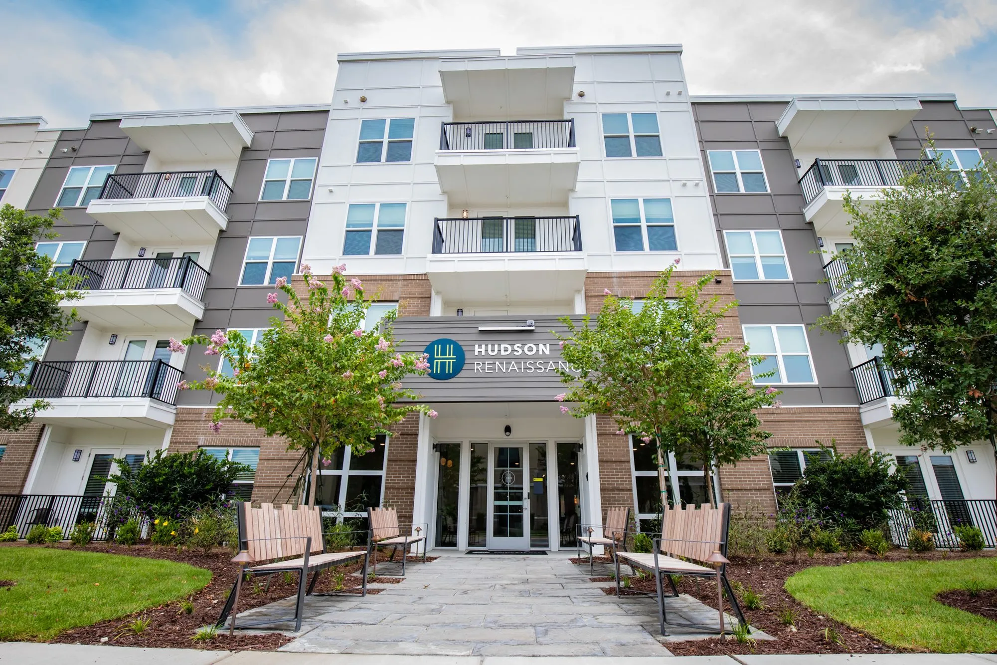 Multifamily community exterior at the front entrance of the four storey building, slatted benches between flowering trees