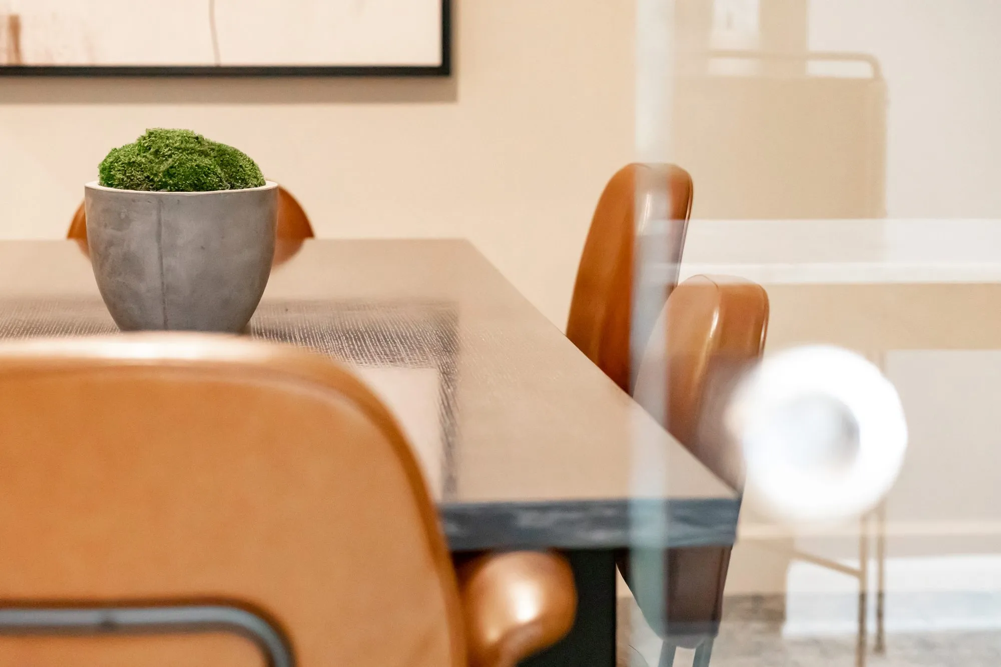 Multifamily amenity interior detail: tan leather chairs at a dark table with a moss ball planter, seen through a glass wall
