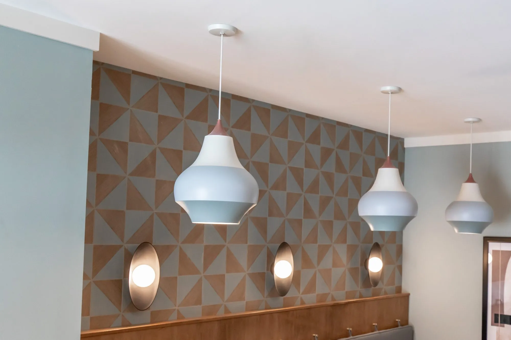 Multifamily amenity lobby with white bell pendants and oval sconces above a wood bench against geometric wallcovering