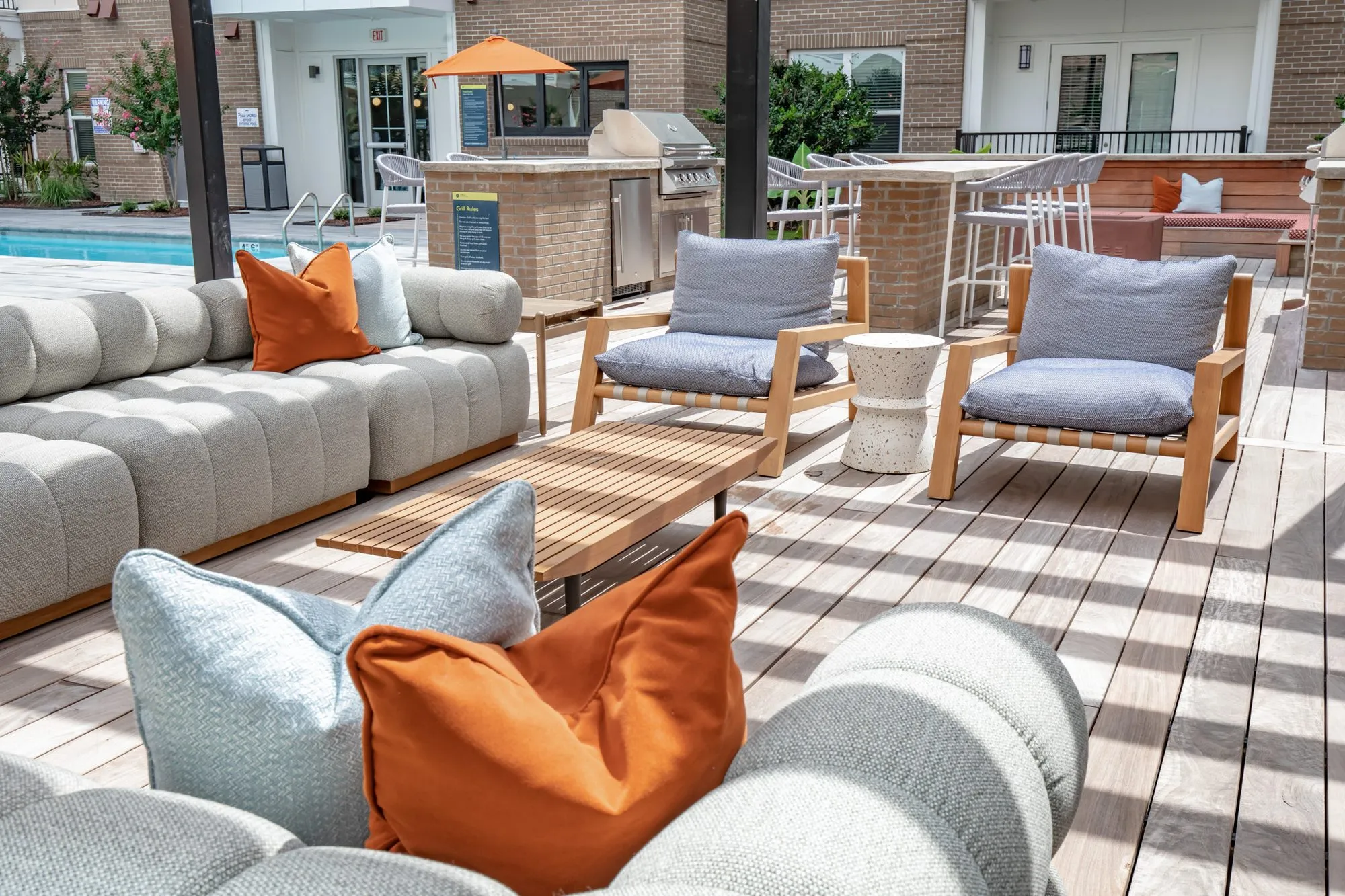 Multifamily community pool deck seating of grey channel-tufted sofas and teak-framed chairs, the outdoor bar beyond