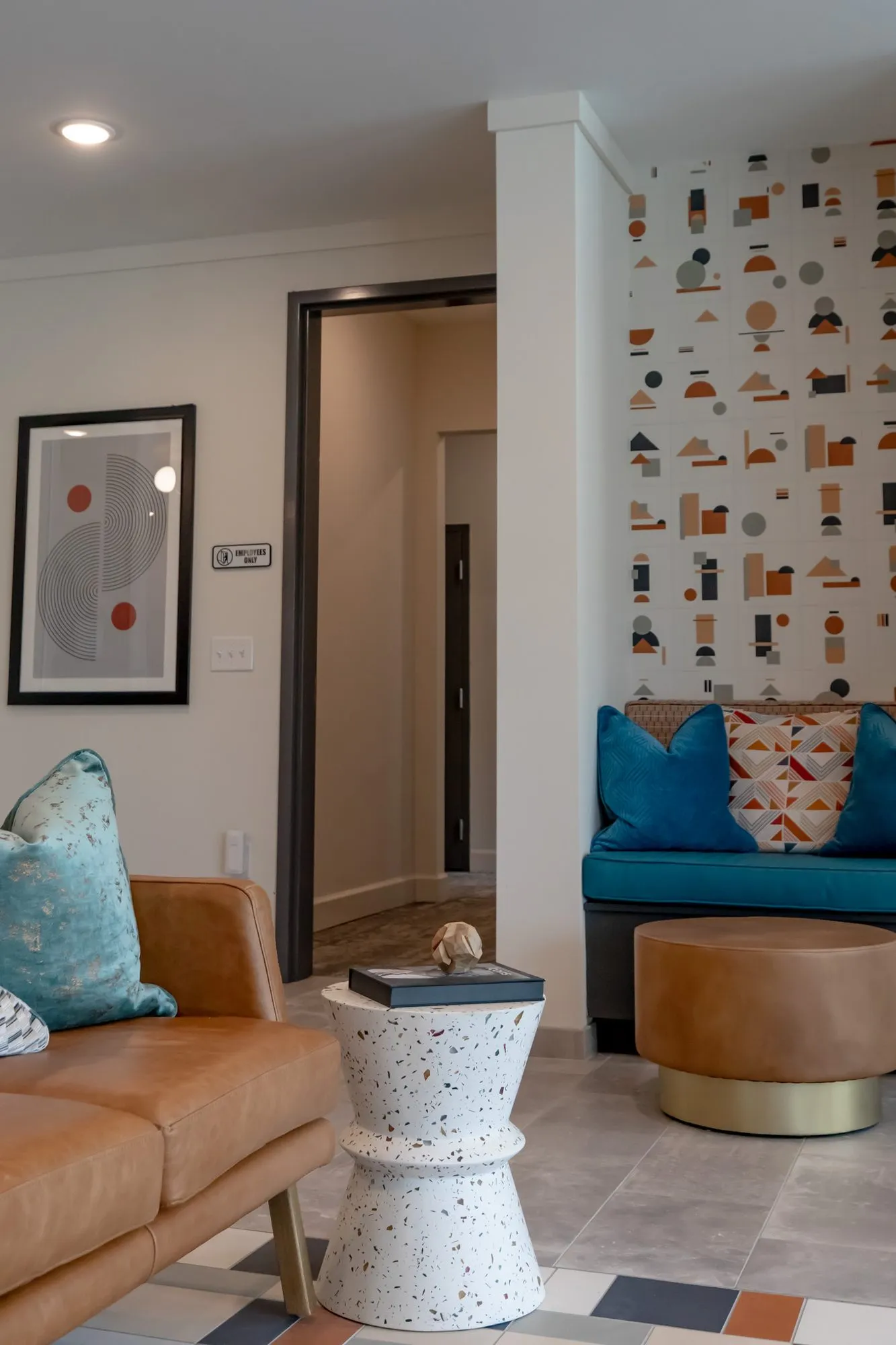 Multifamily amenity lobby lounge with a tan leather sofa and terrazzo side table near a teal bench nook and a framed print