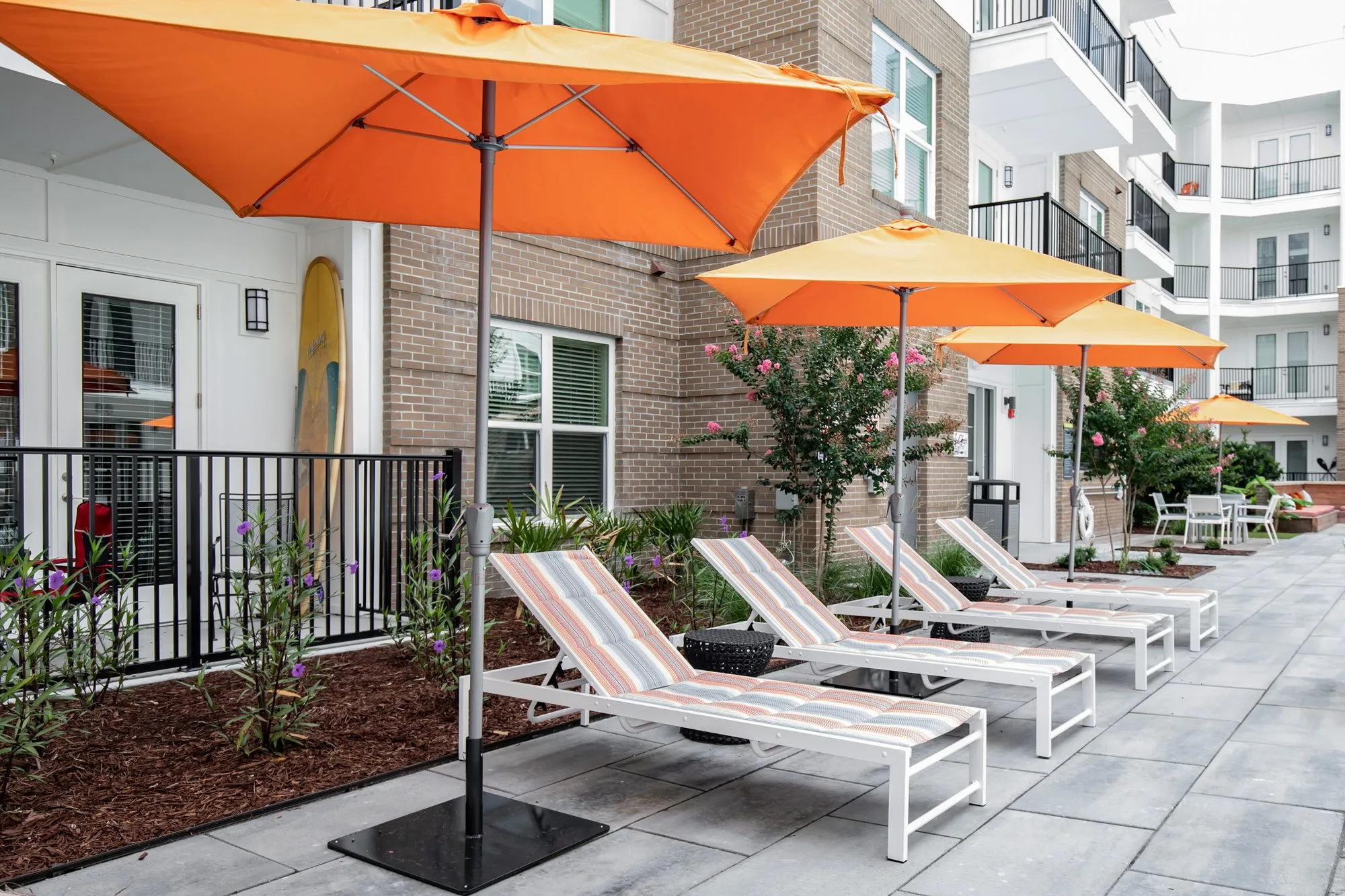 Multifamily community exterior: ground floor patios with striped chaise loungers under orange umbrellas along a paved walk