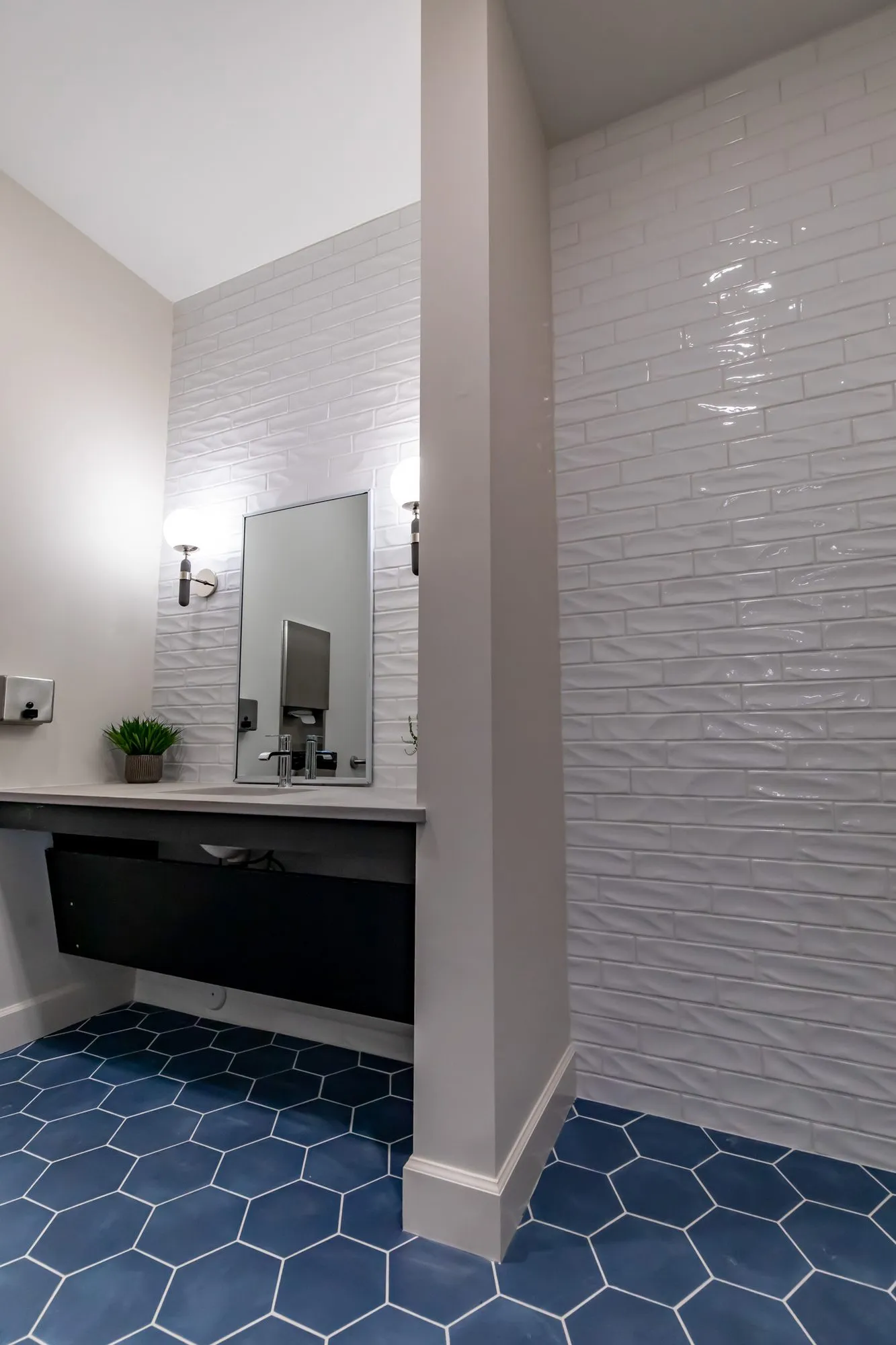 Multifamily amenity restroom seen past a glossy white wave tile wall to the floating vanity, navy hexagon tile underfoot